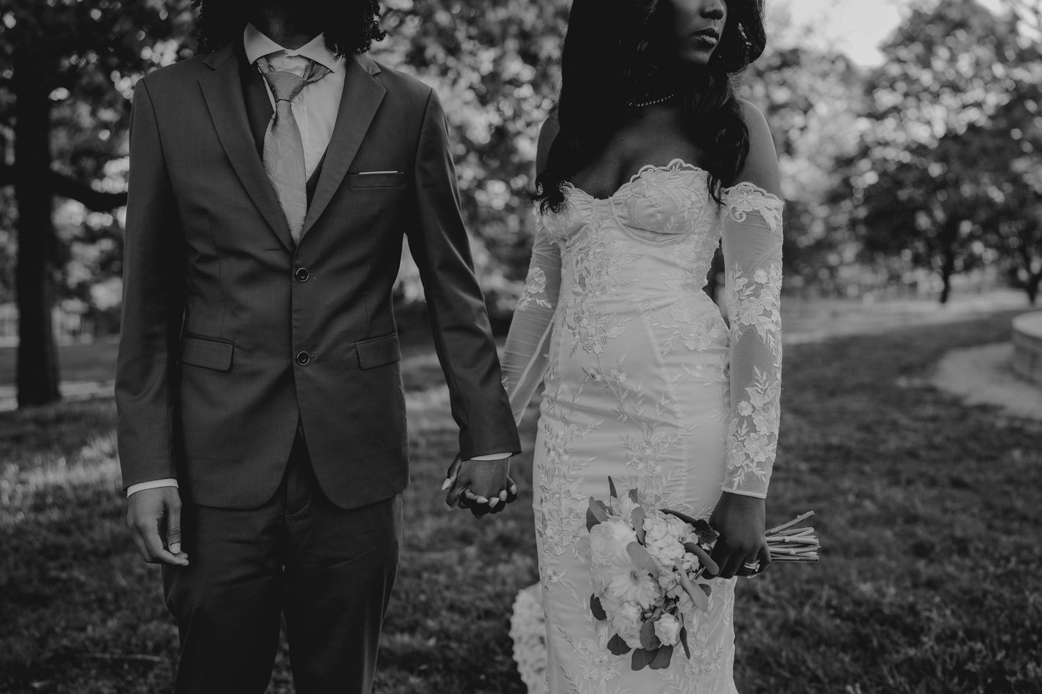 Black and white photo of a bride and groom holding hands outdoors, bride in lace gown holding bouquet.