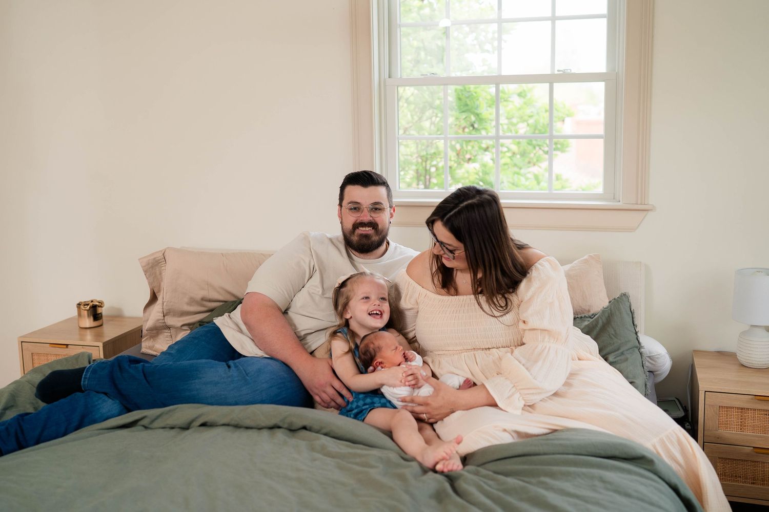 A happy family of three sits on a bed, parents smiling with their young child between them near a bright window.