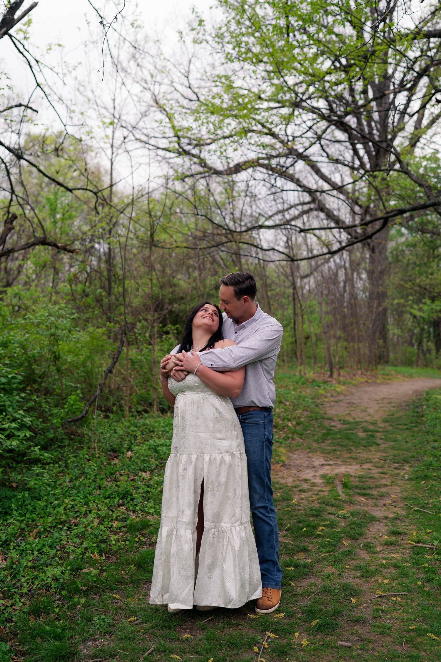 Romantic couple embracing on a forest path in spring, woman in white dress, man in casual attire.