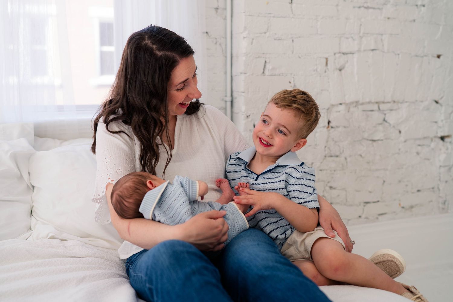 A smiling mother sits on a white bed holding her newborn while her young son laughs beside her.