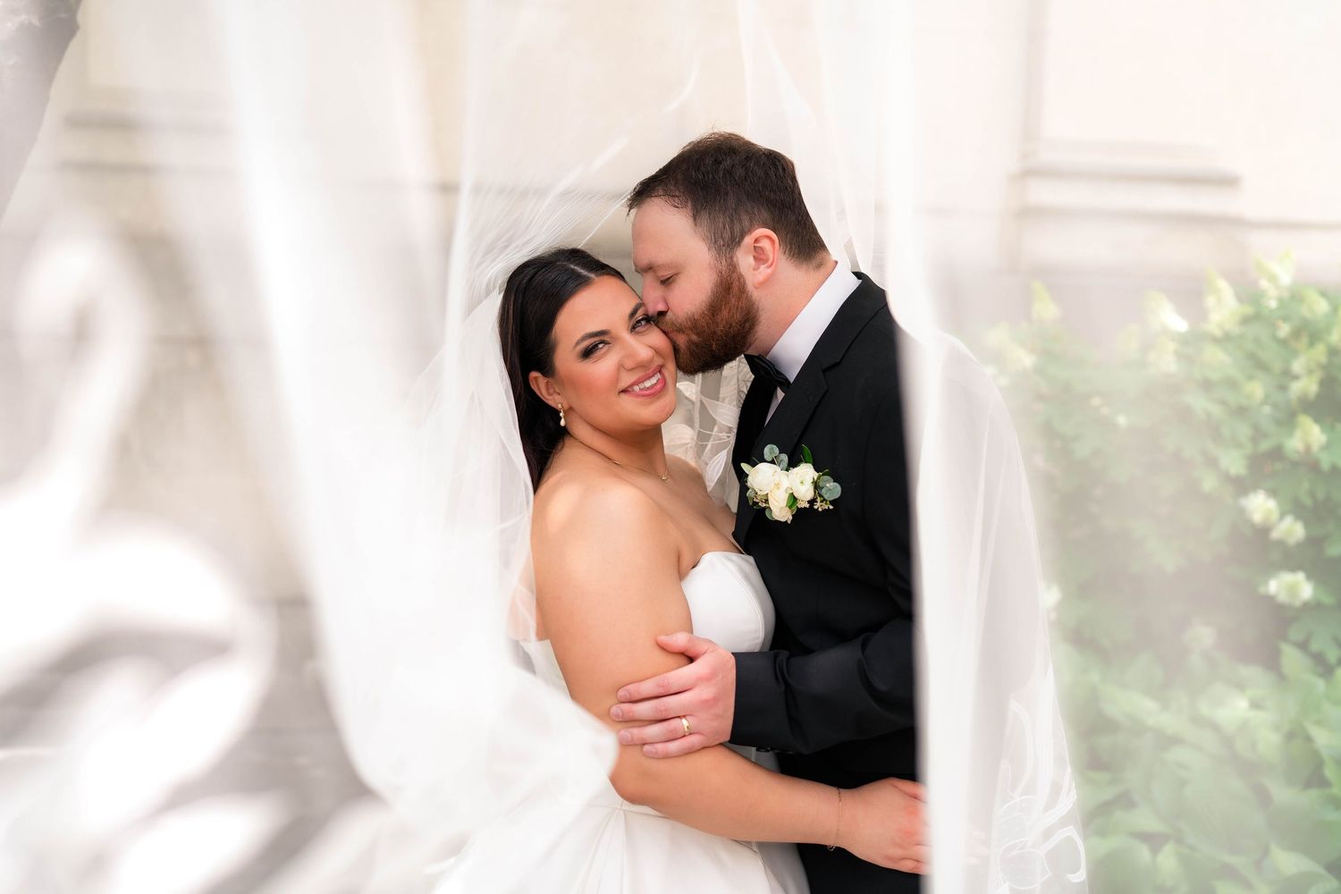 Groom kisses smiling bride on the cheek while wrapped in a flowing white veil.