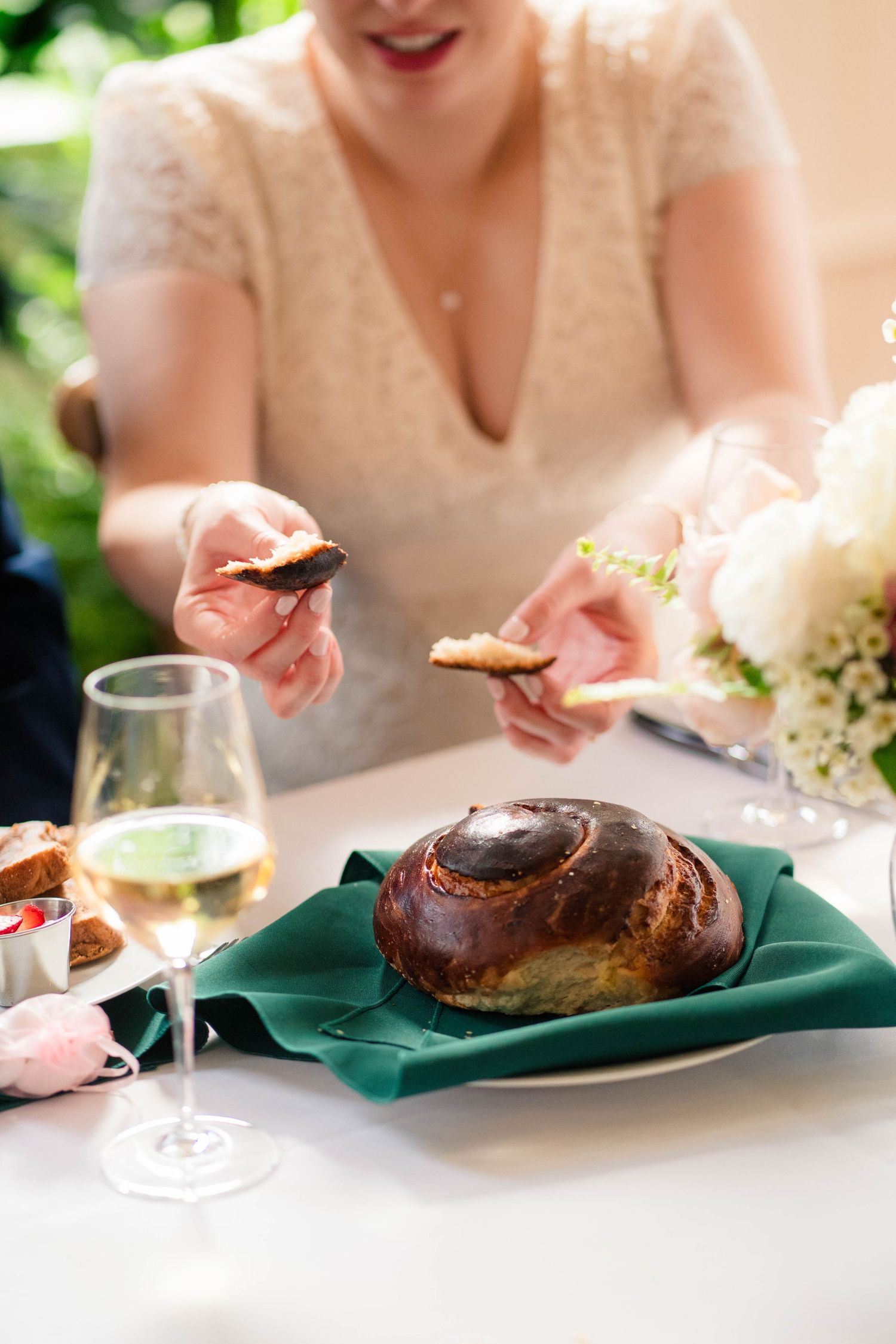 Smiling bride in lace gown breaking challah bread at elegant wedding reception table with wine and florals.