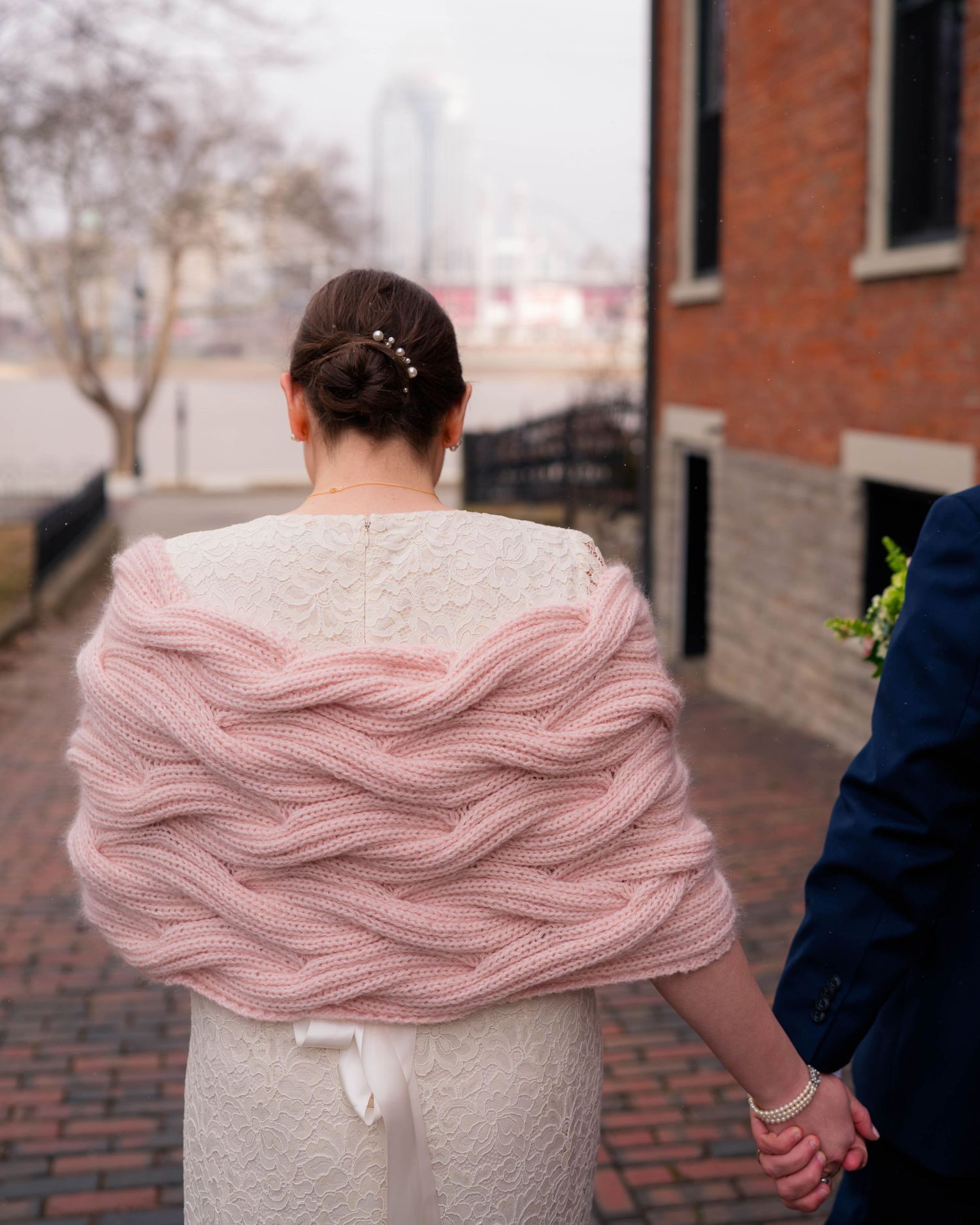 Back view of bride in white lace dress with pink cable-knit wrap shawl on cobblestone street.