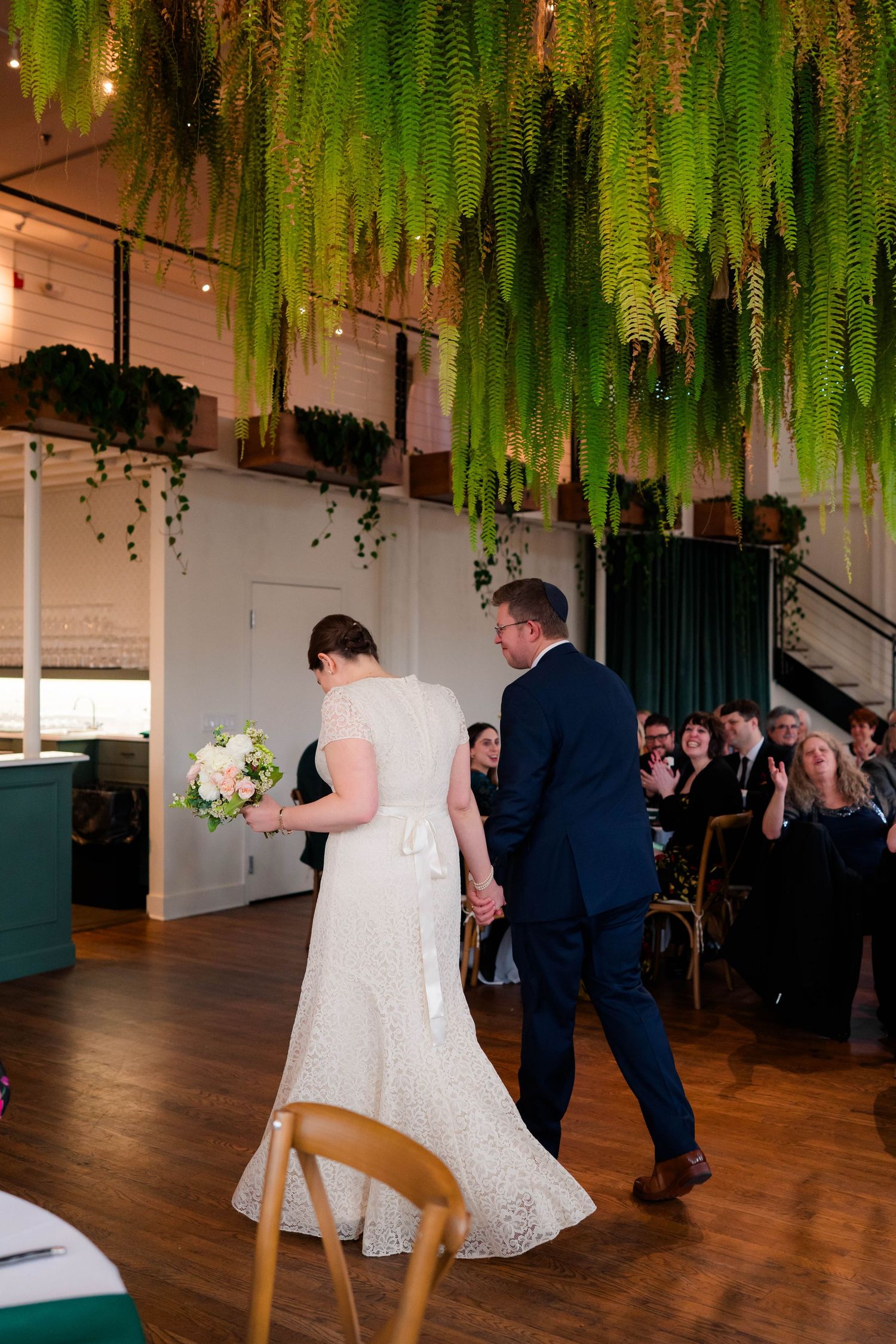 Bride and groom walk hand-in-hand beneath stunning hanging fern canopy at elegant indoor venue.
