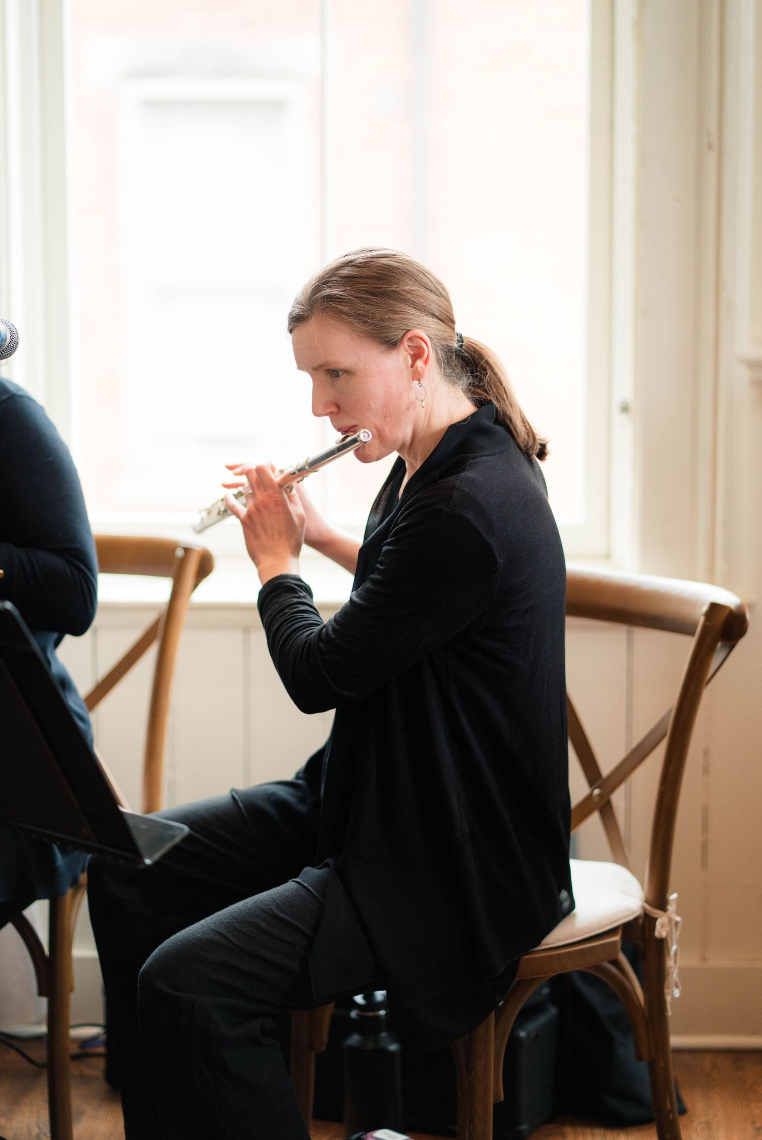 Woman in black outfit plays silver flute while seated at a formal indoor musical performance.