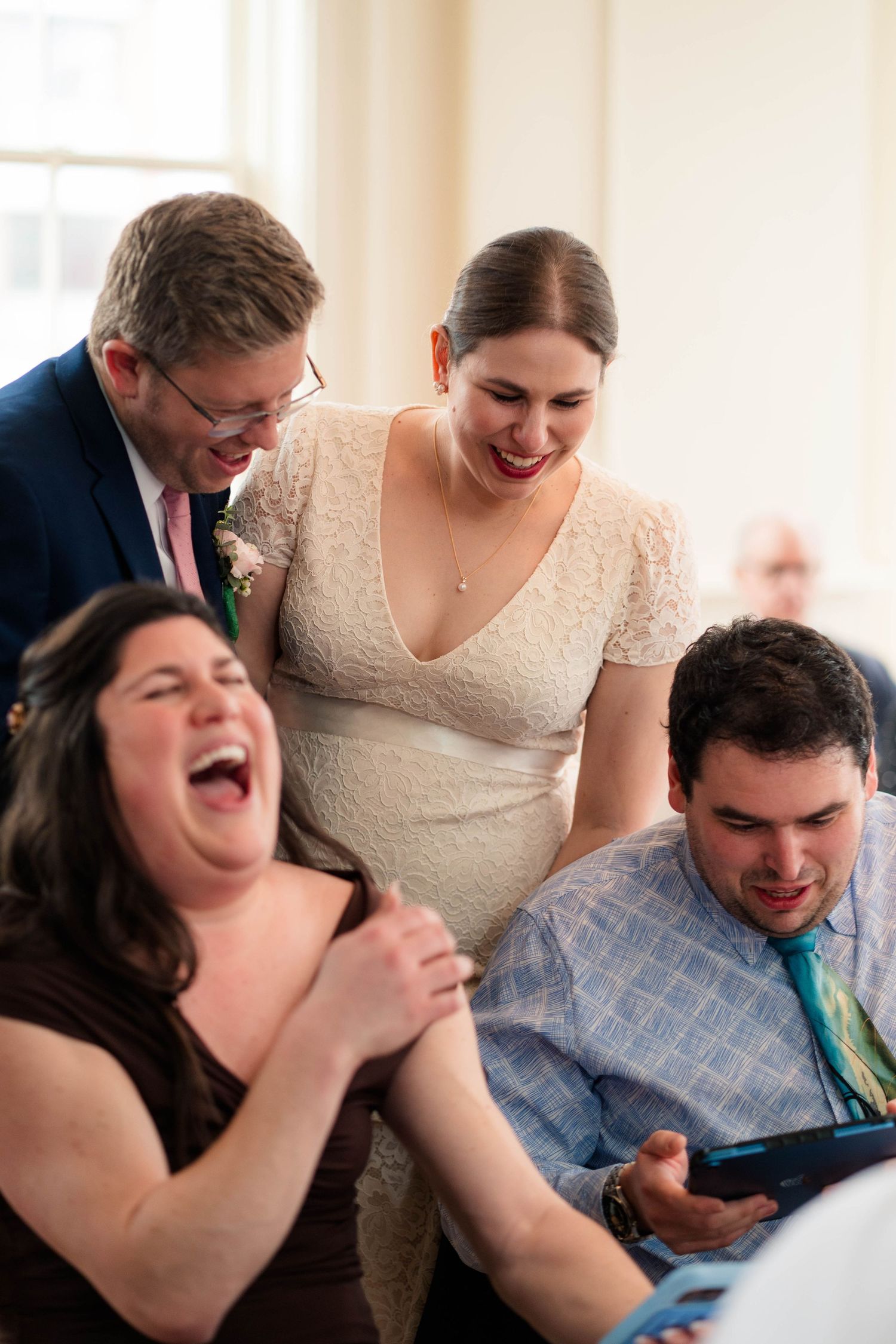 Bride and groom sharing laughs with guests viewing tablet at lively wedding reception celebration.