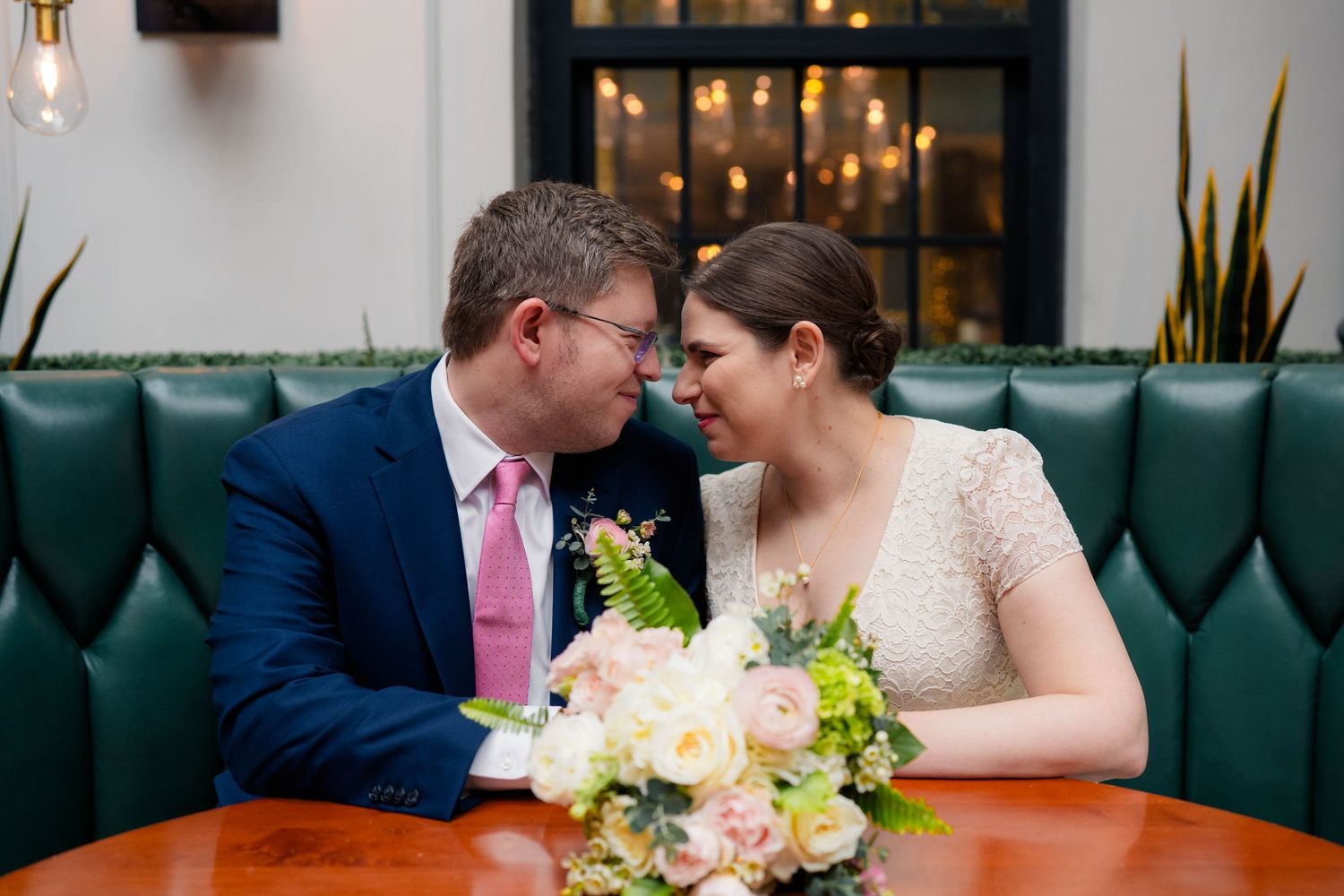 Newlyweds lean in for a romantic nose-to-nose moment seated at a green velvet restaurant booth.
