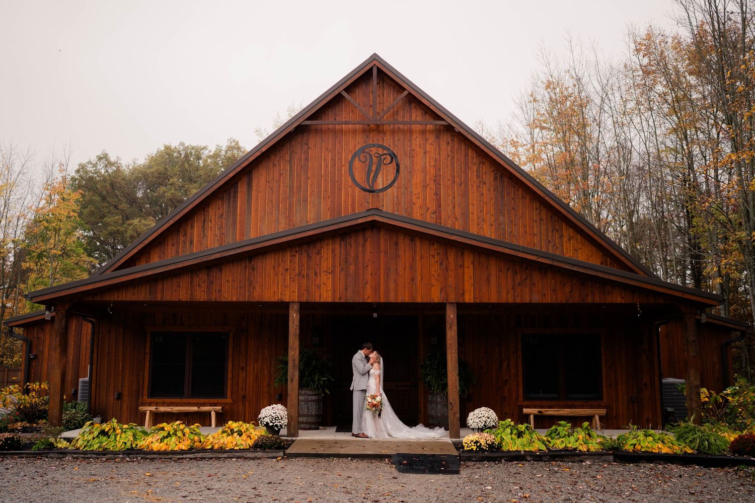 Bride and groom kiss in front of a large rustic wooden barn wedding venue on a misty autumn day.