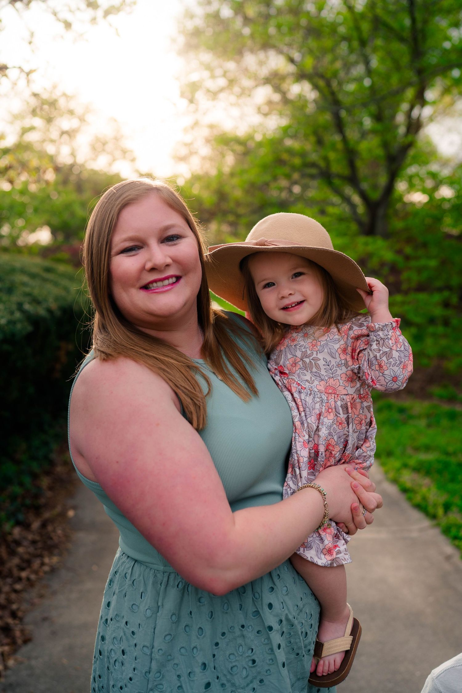 A smiling woman in a teal dress holds a young girl wearing a floral dress and wide-brim sun hat outdoors.