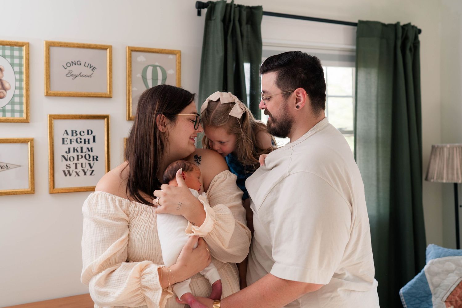 Young family of four shares tender moment, mother holding newborn while toddler daughter kisses baby.