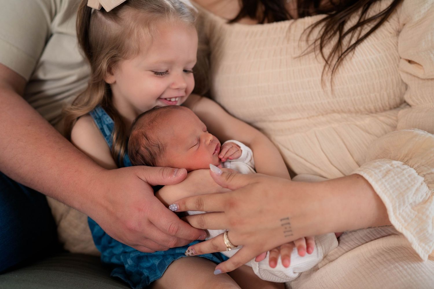 A toddler girl smiles at a newborn baby held by parents during a family newborn photography session.