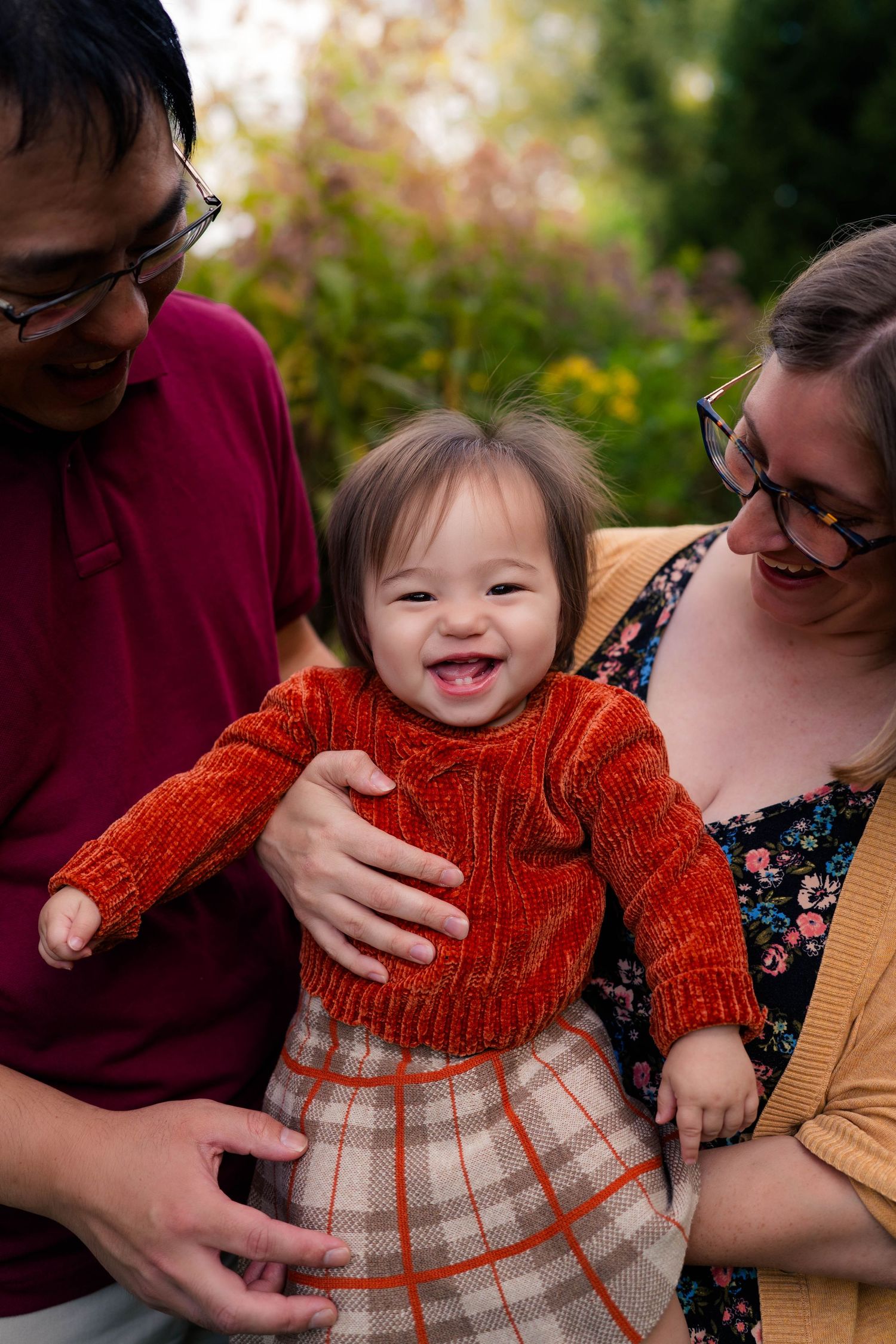 Joyful toddler in red sweater laughing while being held by parents outdoors in autumn garden.