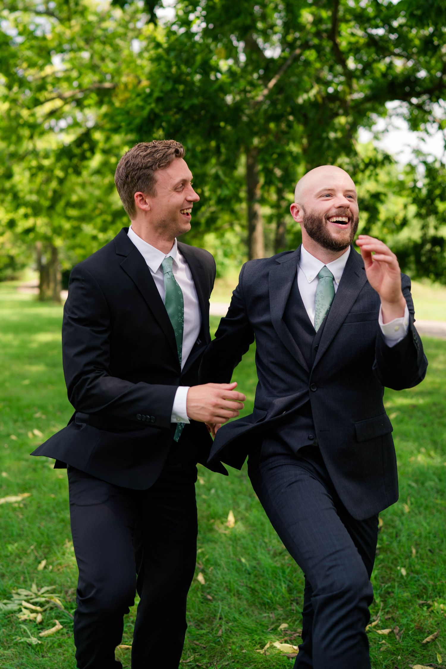 Two groomsmen in dark suits with green ties laugh together outdoors at a wedding celebration.
