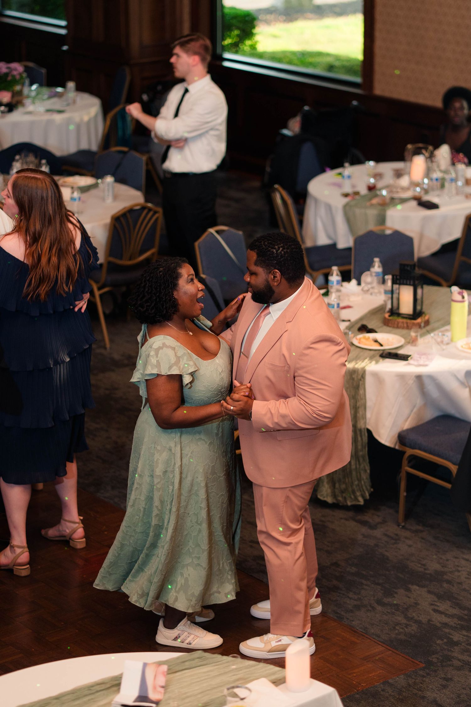 Couple in sage green dress and blush pink suit share a joyful dance at wedding reception.