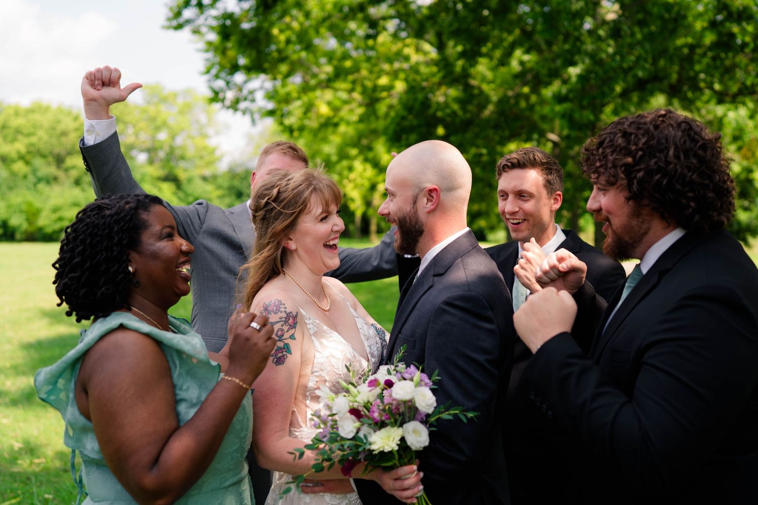 A joyful wedding party celebrates with the bride and groom in a lush outdoor setting.