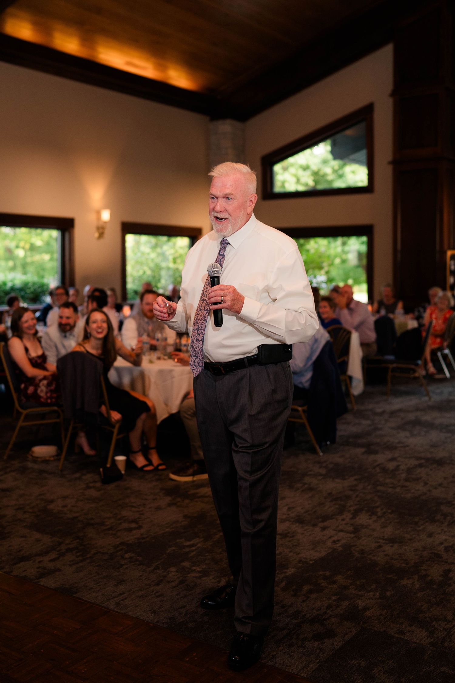 Older gentleman delivers wedding speech holding microphone before seated guests in rustic reception hall.