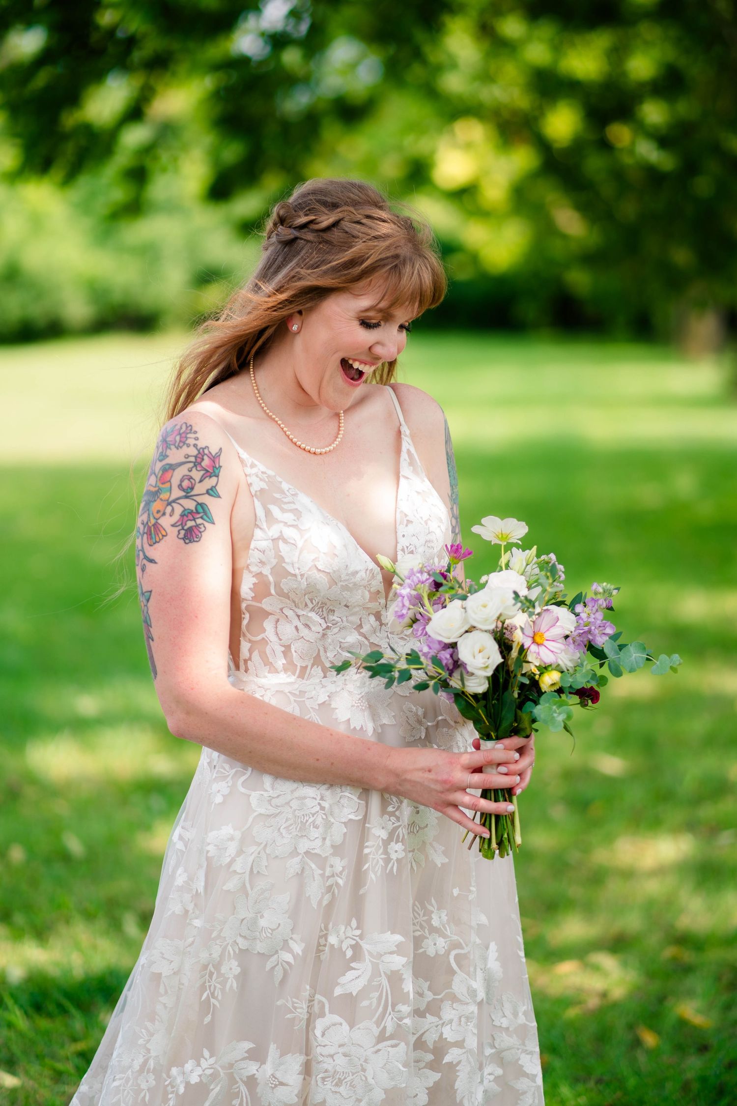A laughing bride in a lace gown holds a purple and white bouquet while standing in a sunny green park.