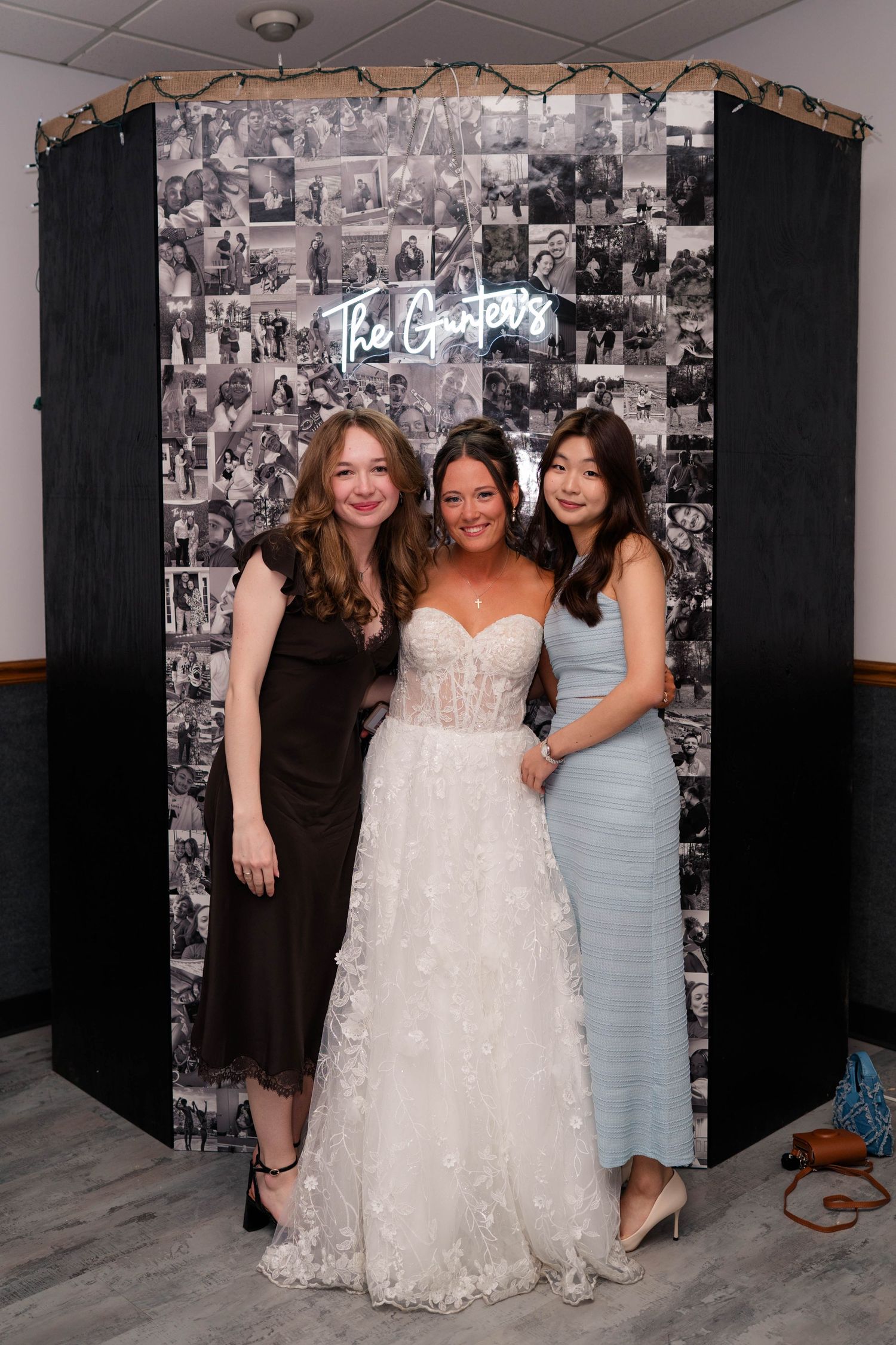 Bride in white lace gown poses with two guests in front of black-and-white photo collage backdrop at wedding reception.