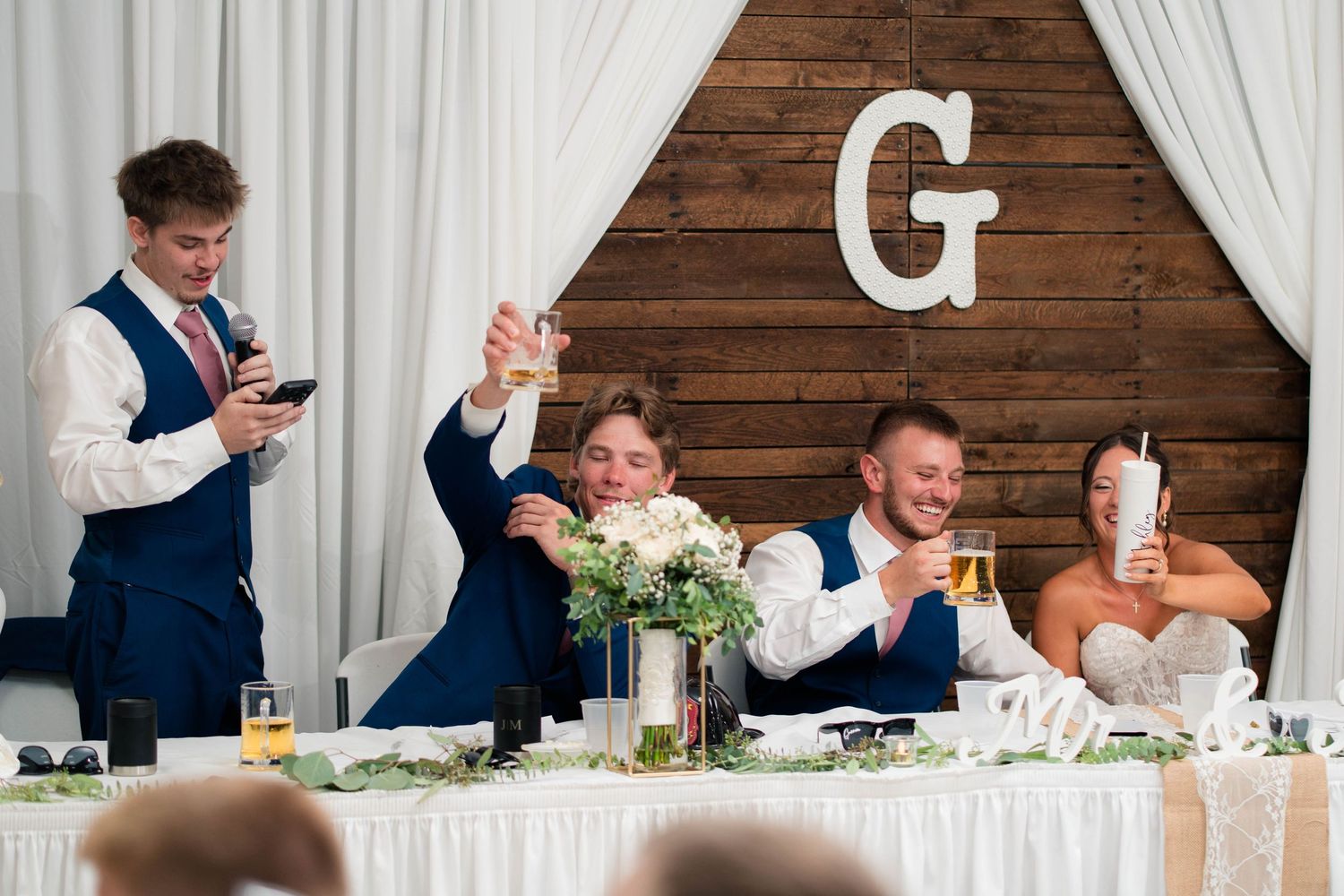 Groom and bride raise glasses in a toast with wedding party at decorated head table during reception.