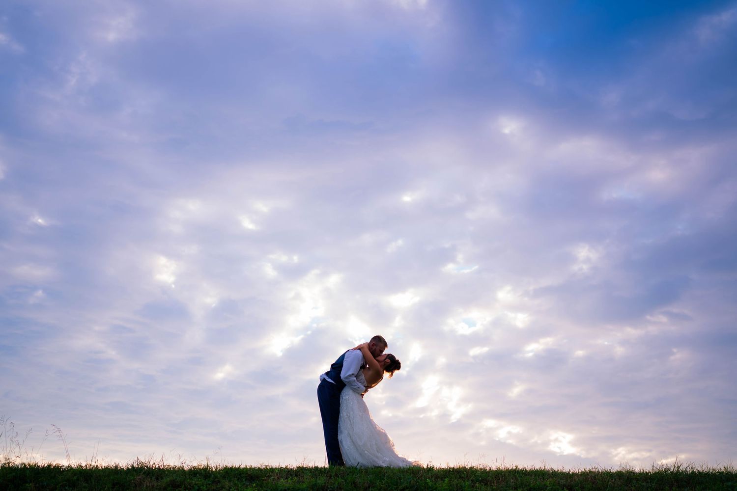 A bride and groom share a romantic dip kiss silhouetted against a dramatic blue cloudy sky at sunset.