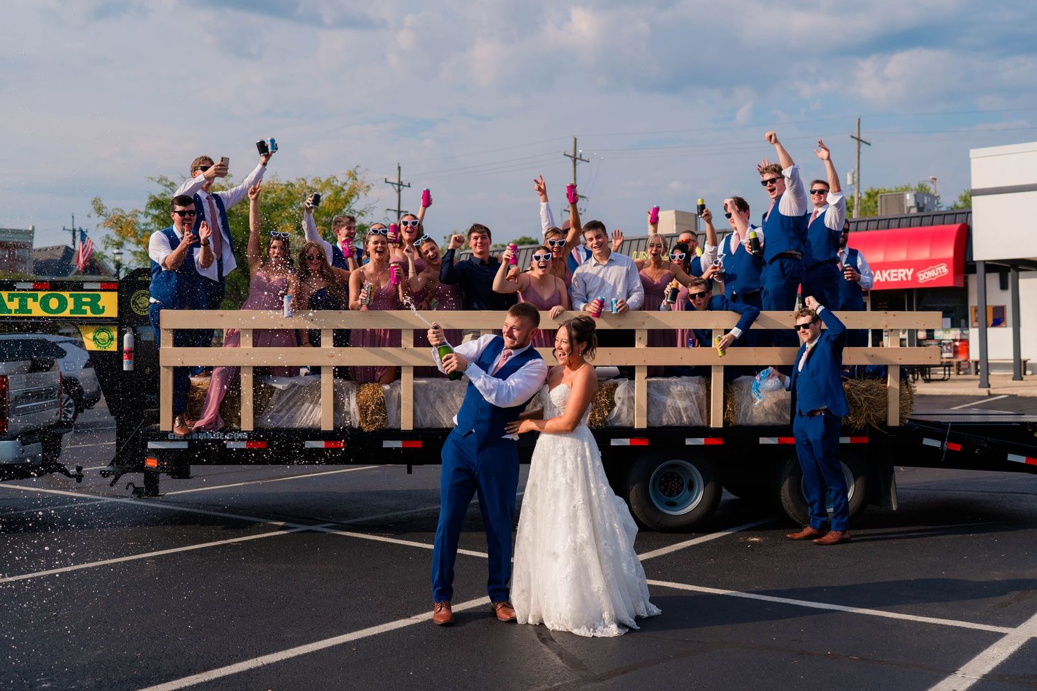 Bride and groom pose in front of a flatbed truck filled with celebrating wedding guests in a parking lot.