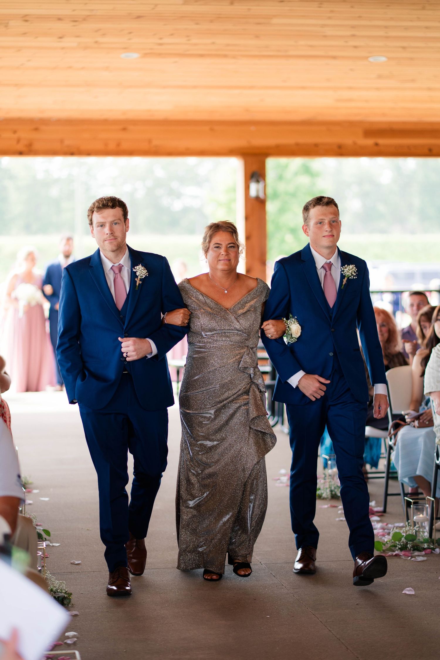 Woman in silver gown walks down wedding aisle escorted by two men in navy blue suits with pink ties.