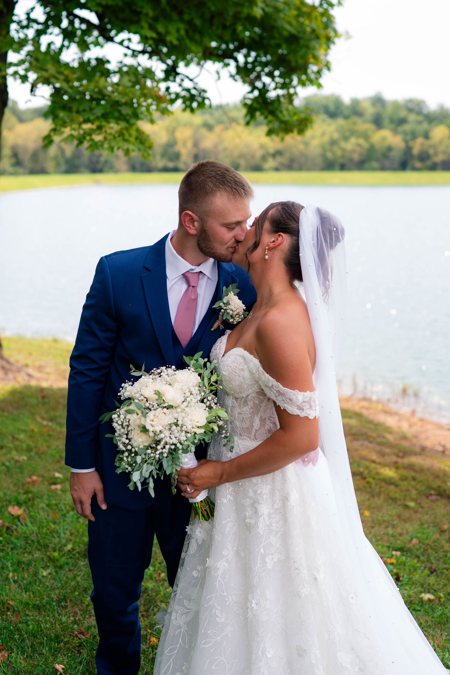 Bride and groom share a romantic kiss near a serene lake, groom in navy suit, bride holding white floral bouquet.