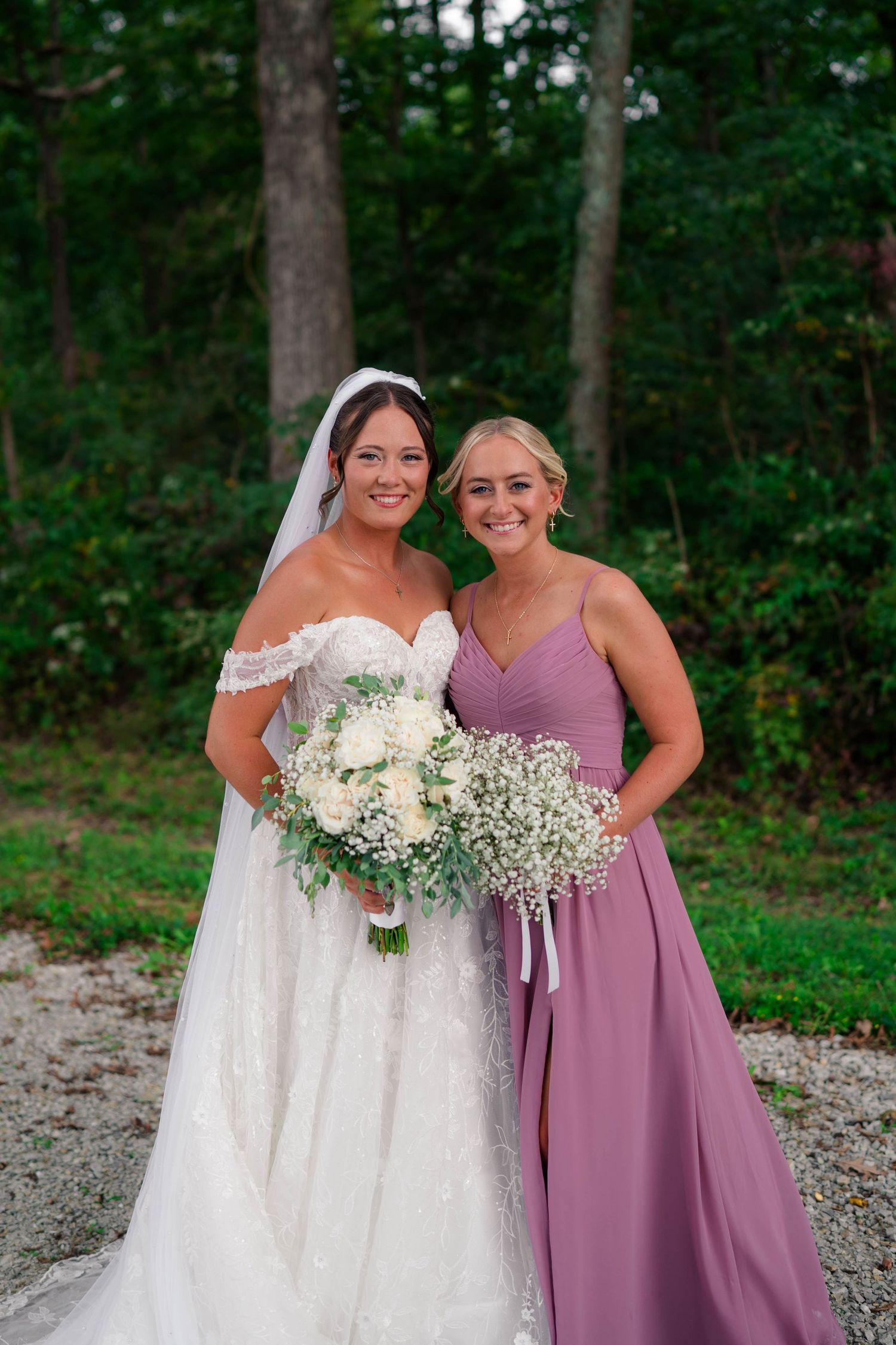 Bride in lace off-shoulder gown poses with bridesmaid in mauve dress holding white floral bouquets.