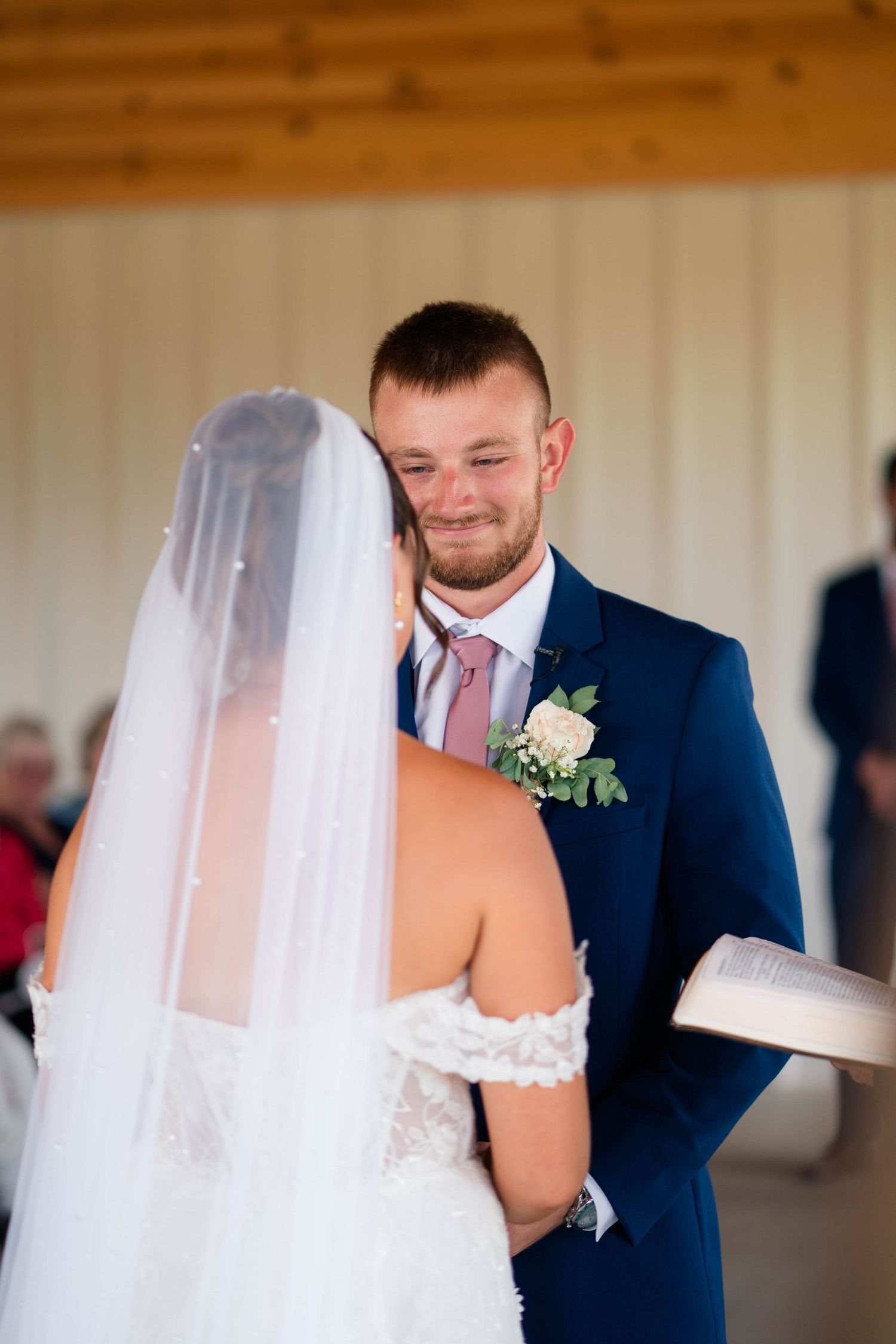 Emotional groom in navy suit smiles during wedding vows while bride in lace veil reads from a book.