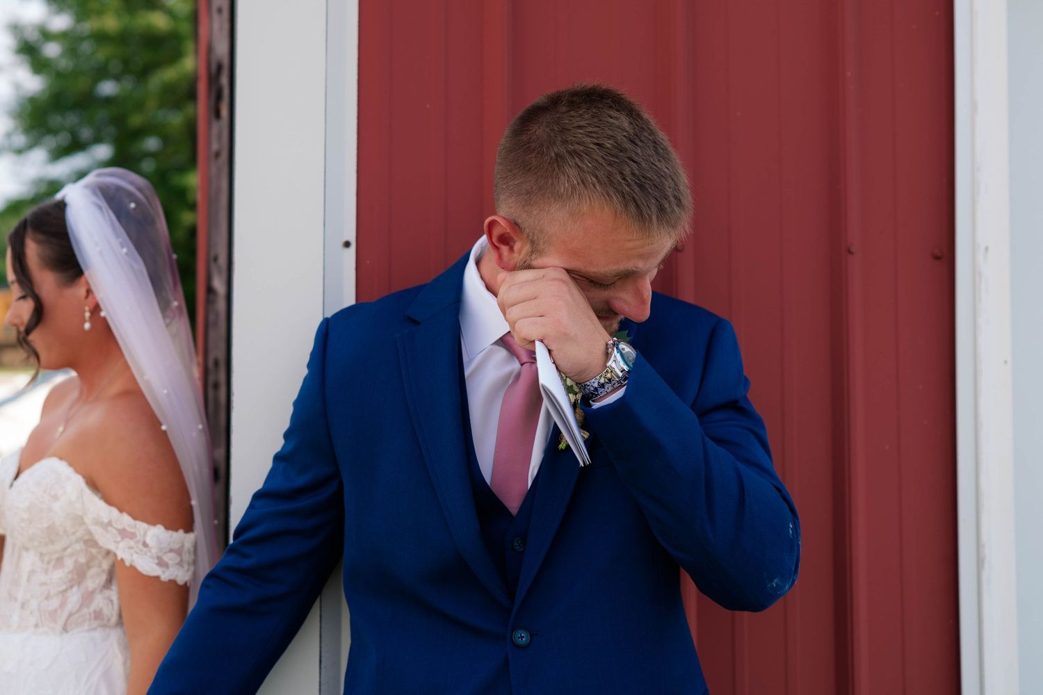 Emotional groom in blue suit wiping tears during first look moment with bride in white dress at barn wedding.