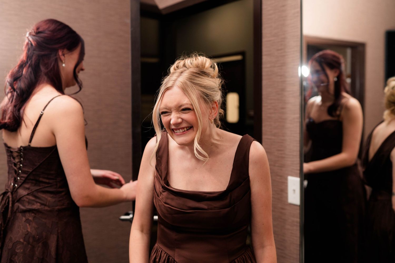 Bridesmaid in brown dress laughing while another bridesmaid helps with her dress.
