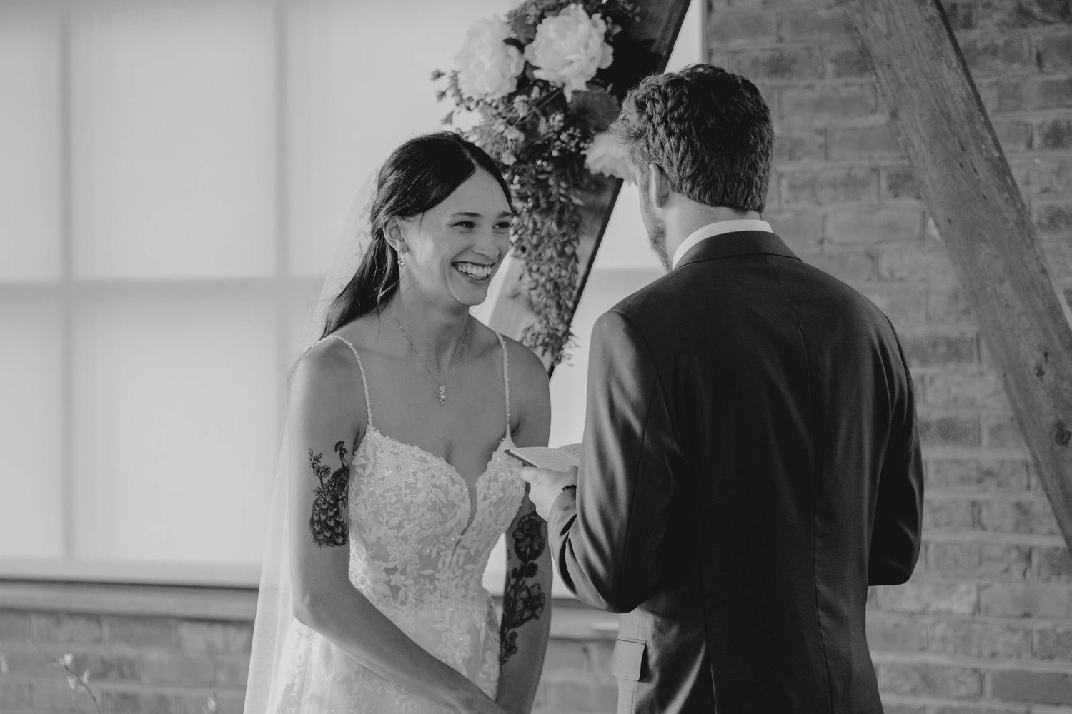 Black and white photo of smiling bride exchanging vows with groom at rustic wedding.