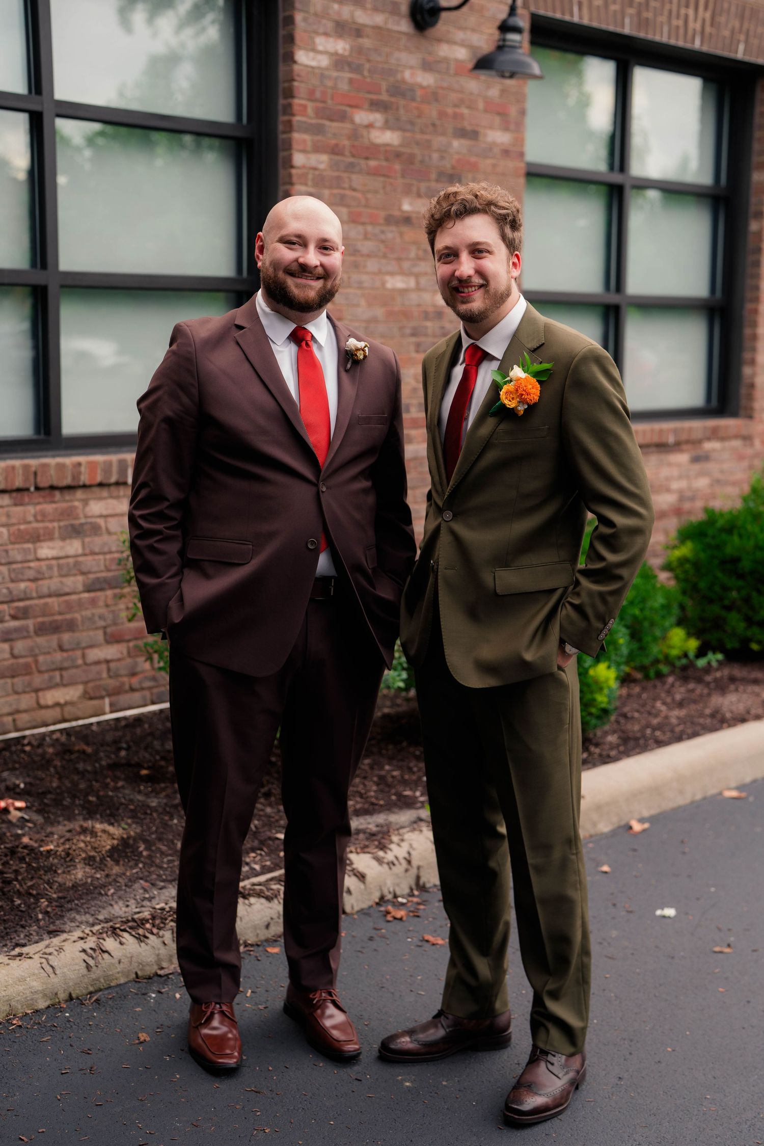 Two grooms in brown and olive suits with red ties and boutonnieres pose outside a brick building.