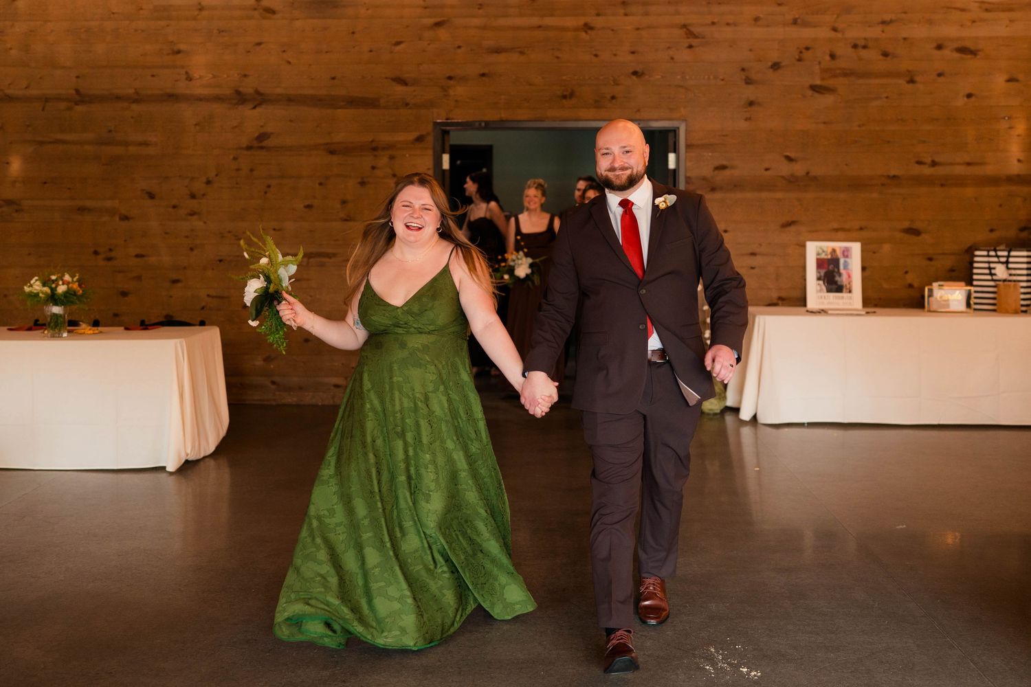 Bridesmaid in emerald green gown and groomsman enter wedding reception holding hands joyfully.