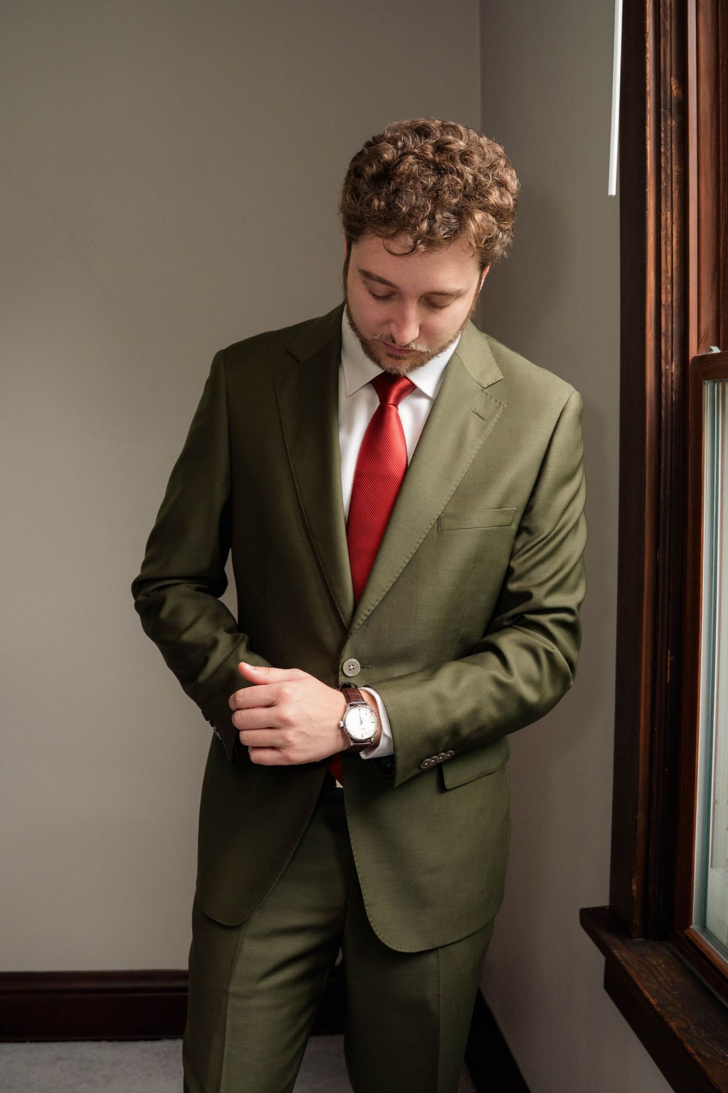 Groom in olive green suit with red tie adjusting his jacket button before wedding ceremony.