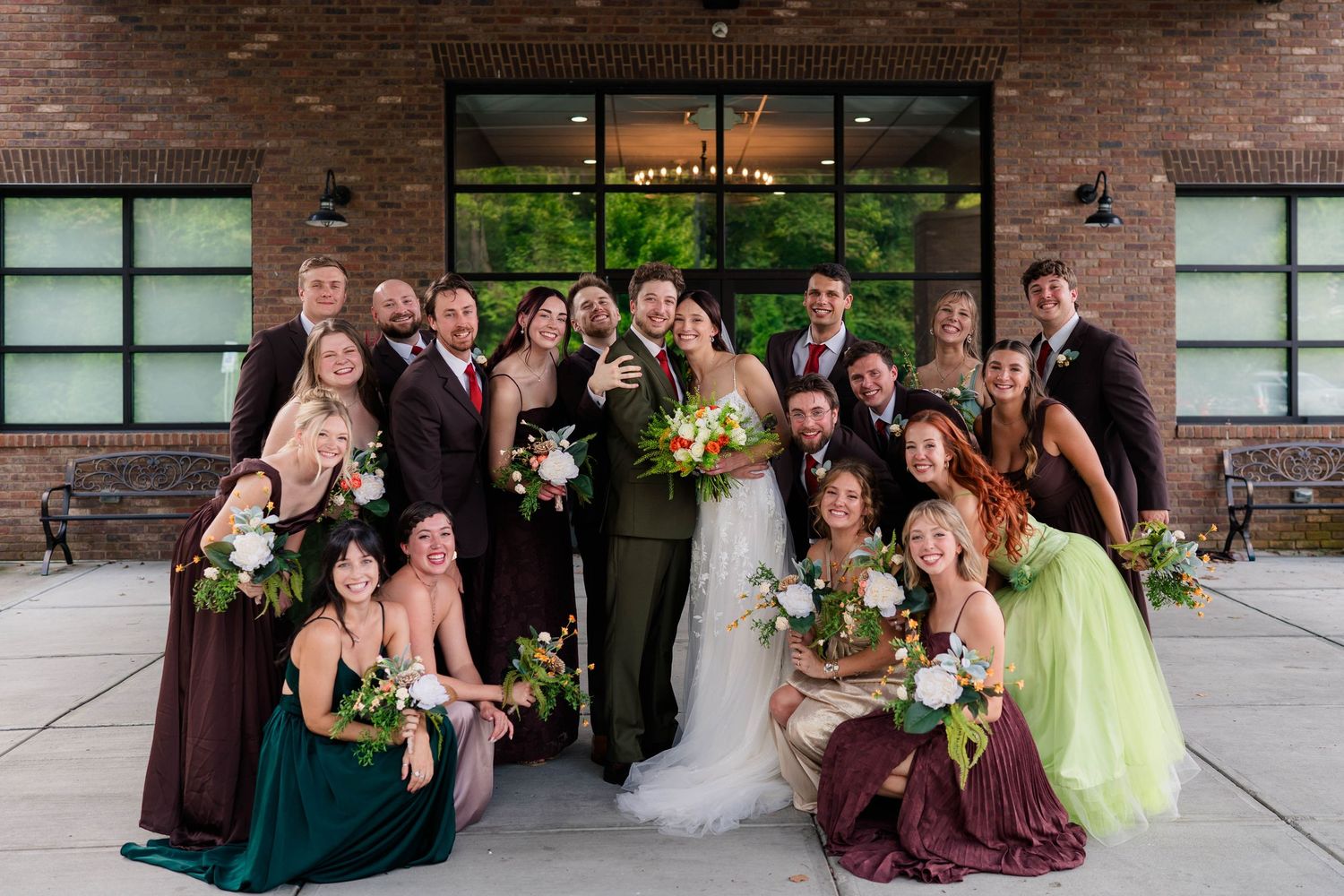 Wedding party group photo with bride, groom, bridesmaids in burgundy and green, and groomsmen outside brick venue.