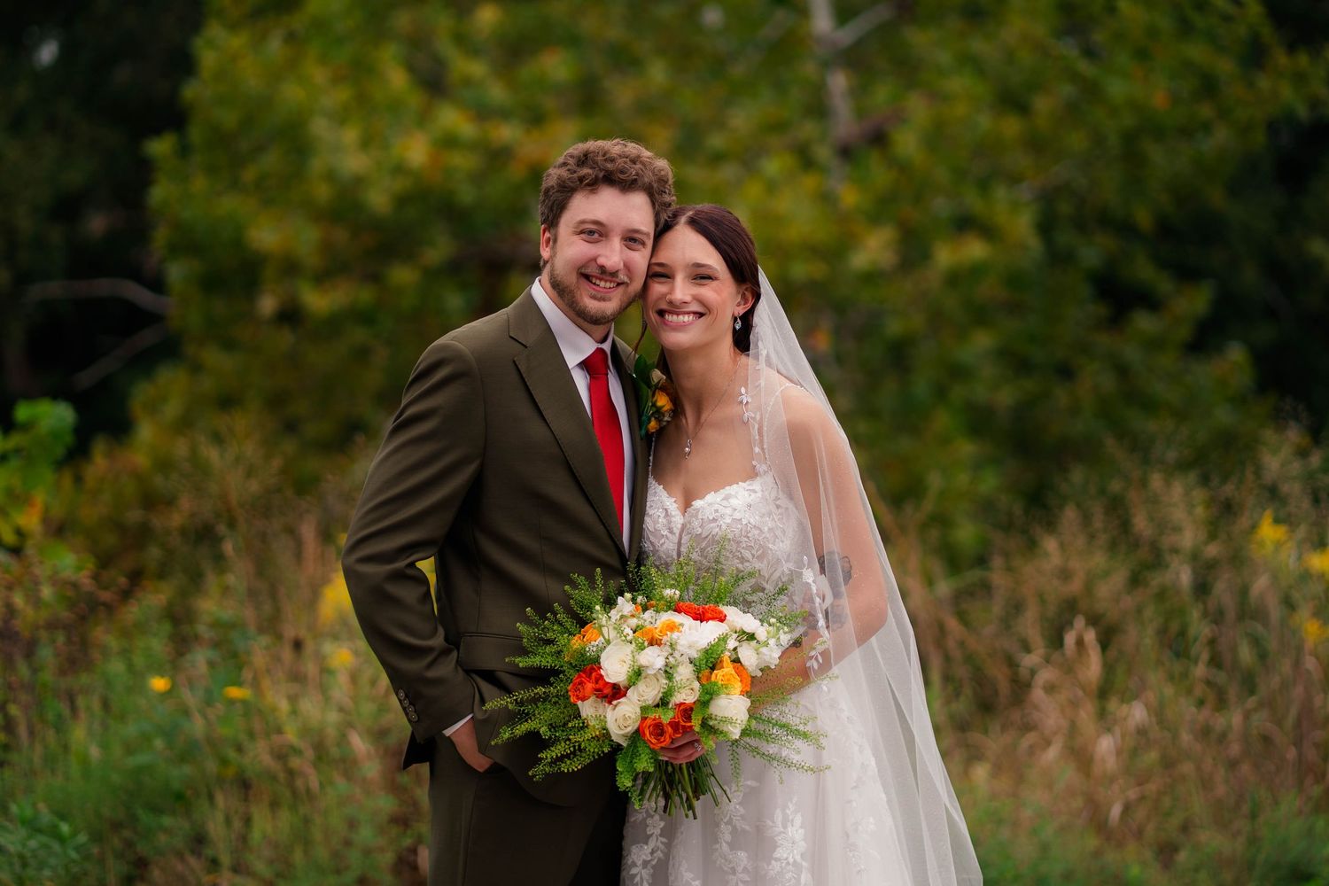 Smiling bride and groom pose outdoors in autumn, bride holding colorful bouquet, groom in olive suit with red tie.