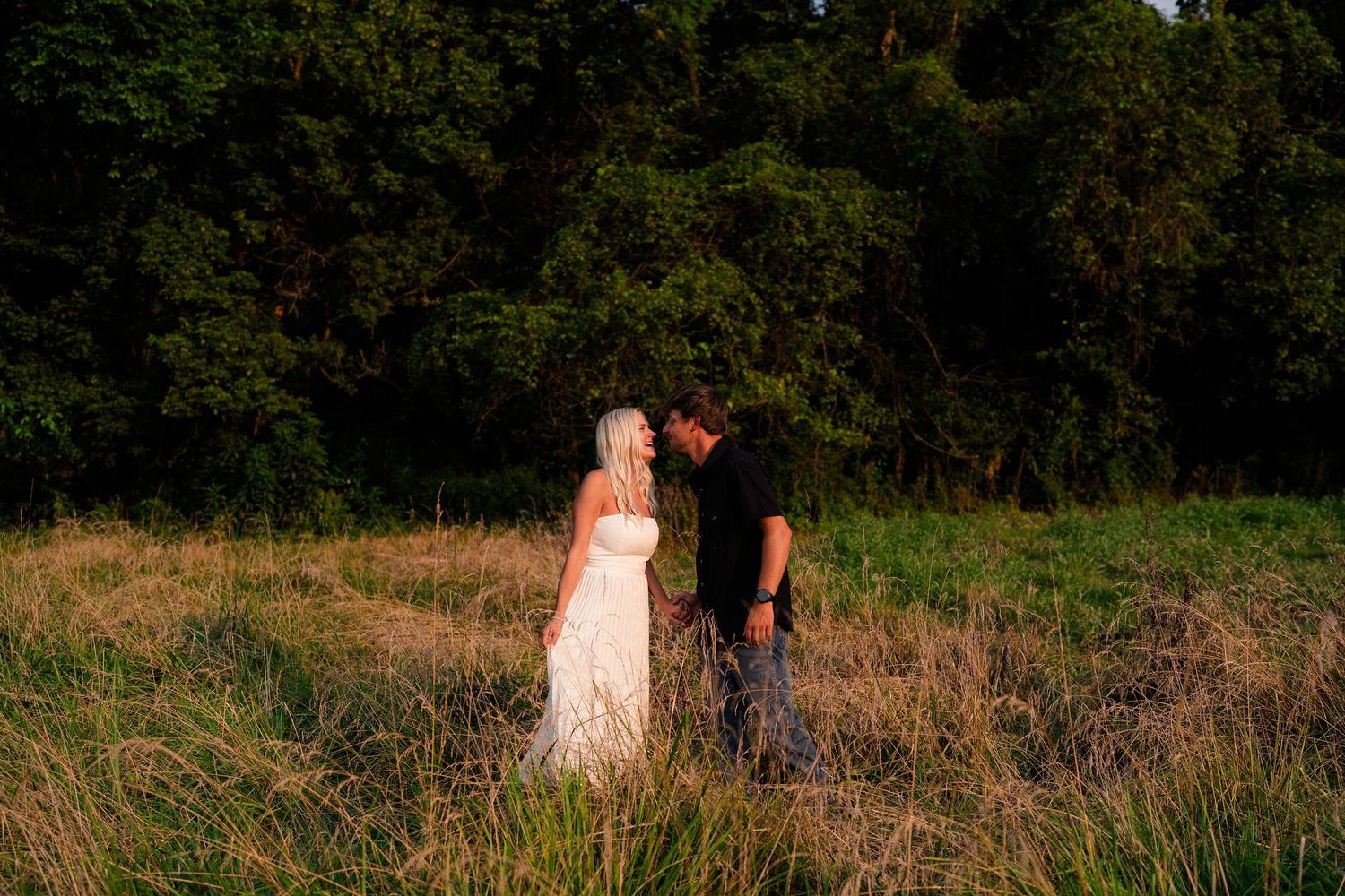 Couple sharing a romantic kiss in a sunlit meadow with lush green trees in the background during golden hour.