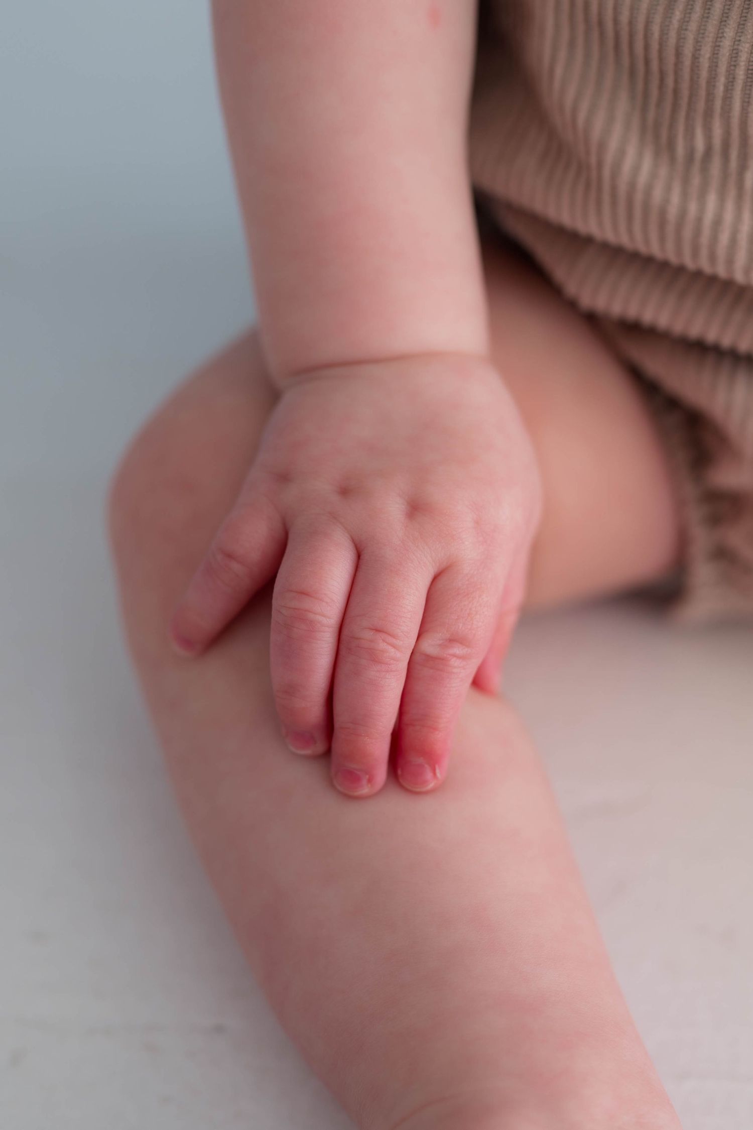 Close-up of a baby's chubby hand resting on their knee, wearing a brown corduroy romper.