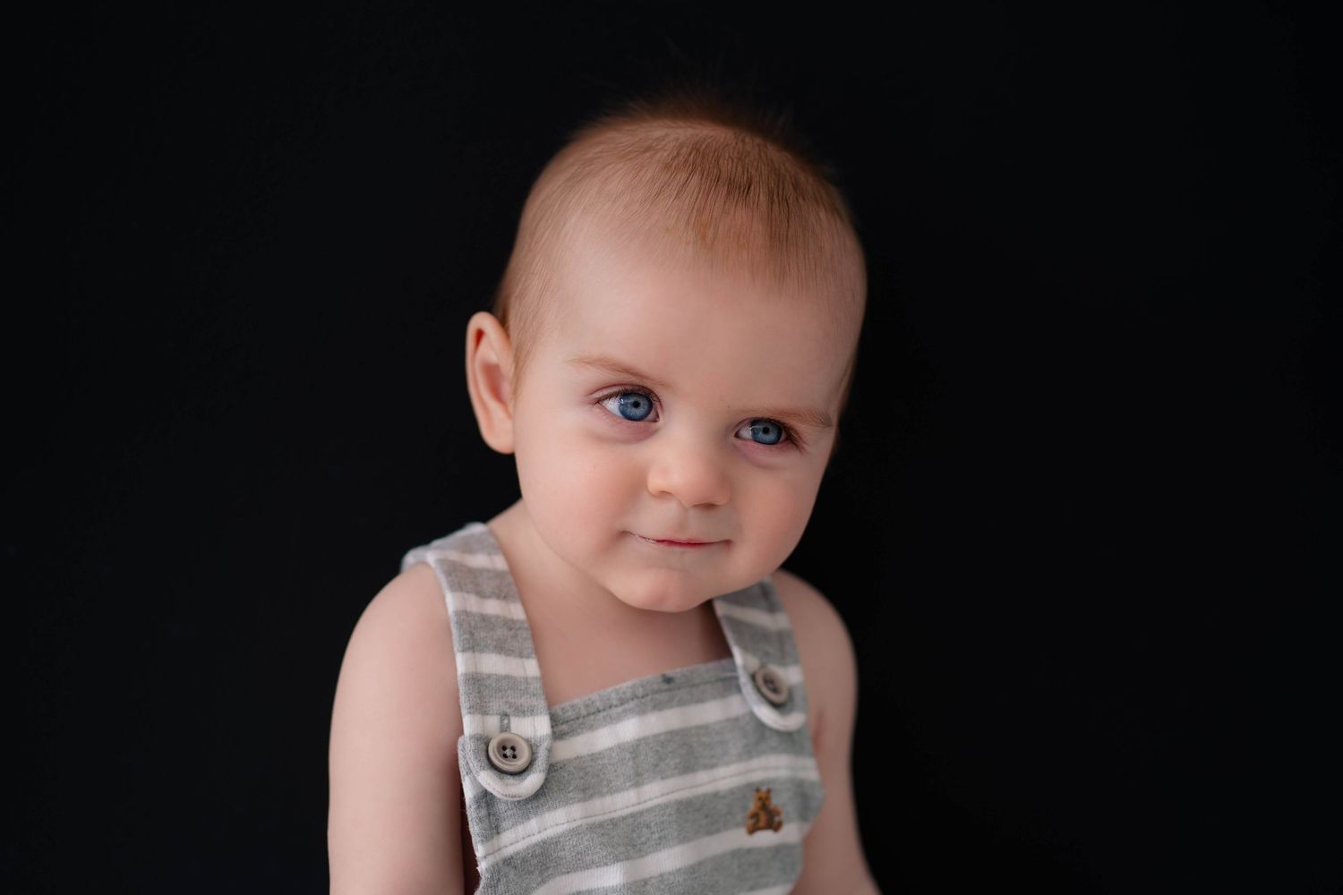 Adorable baby with blue eyes wearing striped overalls poses against a black background in a professional portrait.