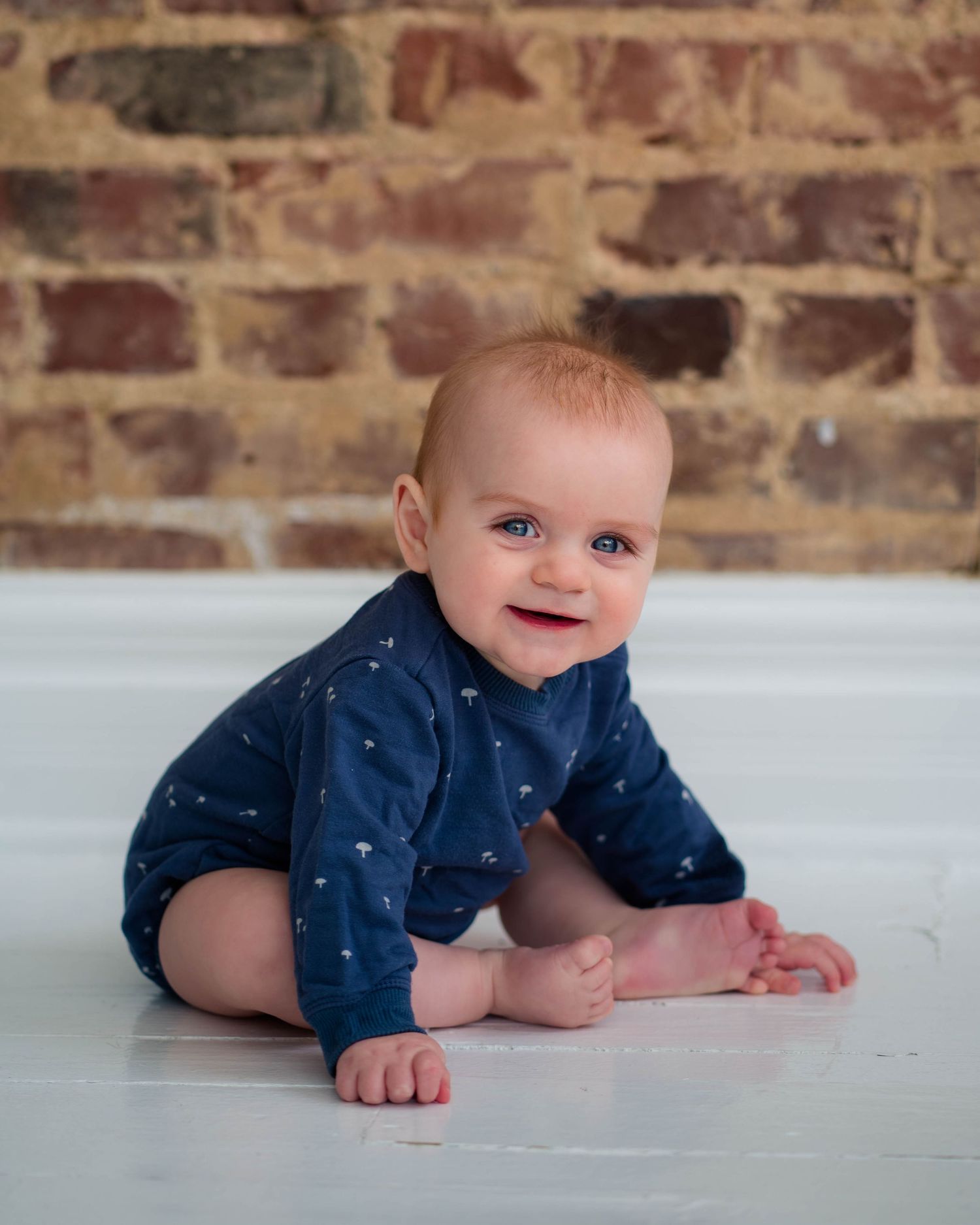 Smiling blue-eyed baby in navy romper crawling on white floor with brick wall background.