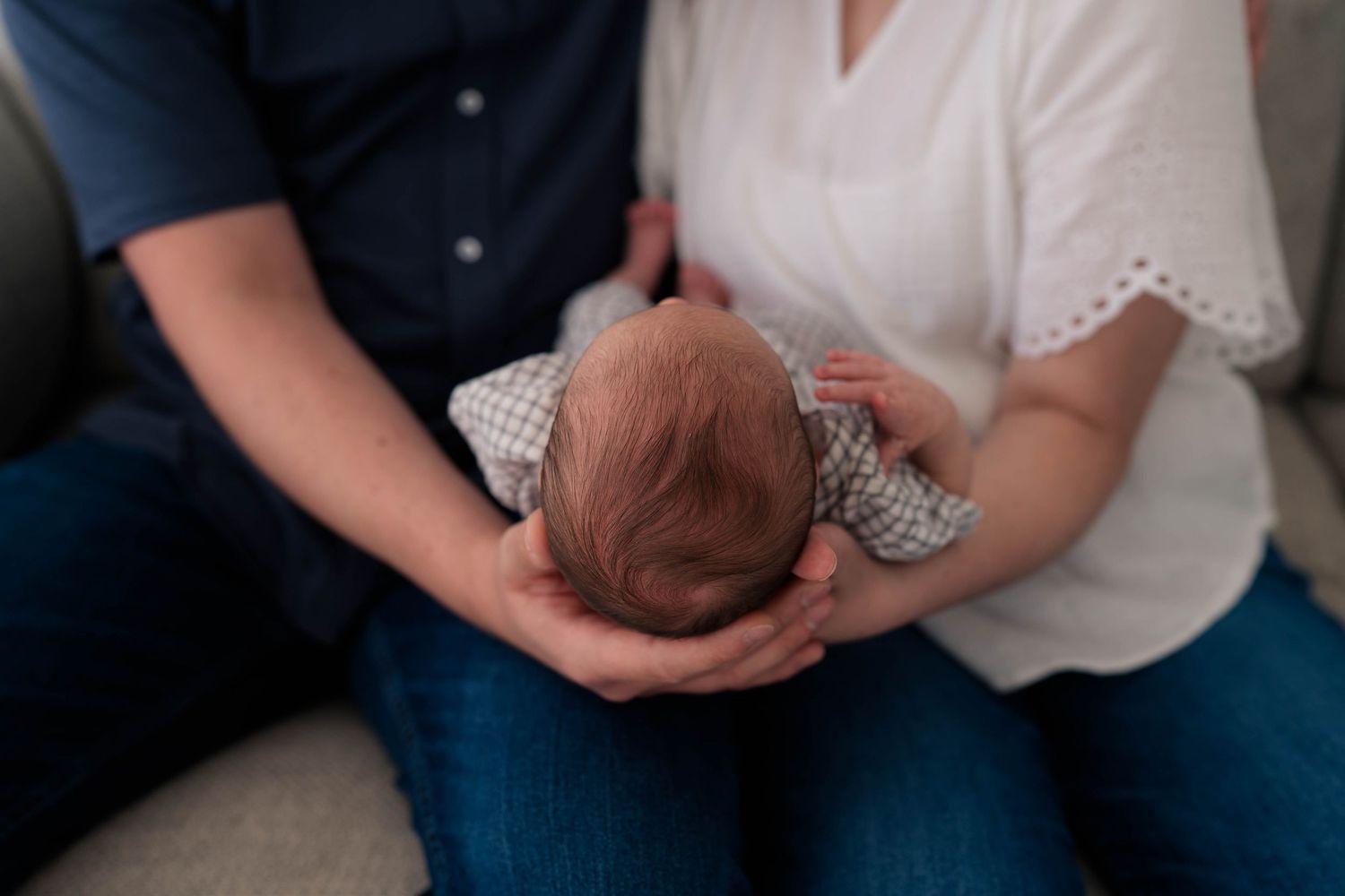 Two parents gently cradle their newborn baby together, showing loving hands supporting the infant dressed in a patterned outfit.