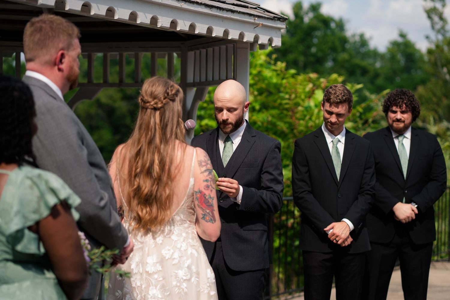 Bald groom places ring on brides finger during outdoor wedding ceremony with groomsmen in black suits behind.