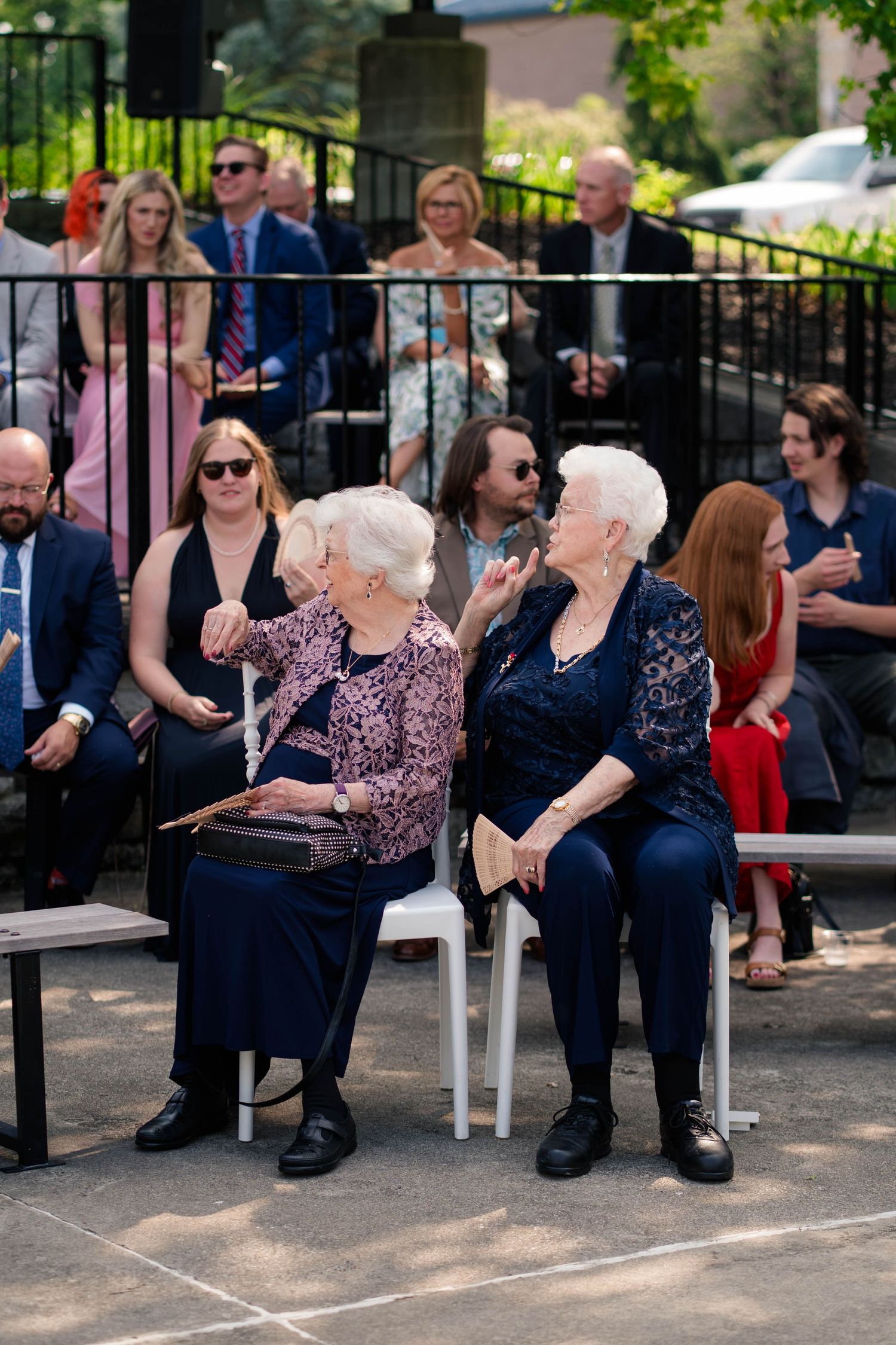 Two elderly women chat and laugh together while seated at an outdoor wedding ceremony with guests in background.