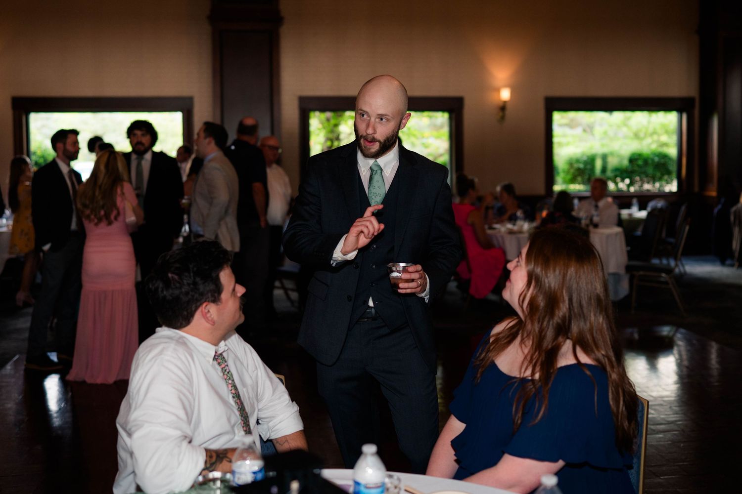 Bald groom in dark suit mingles with wedding reception guests at elegant venue.