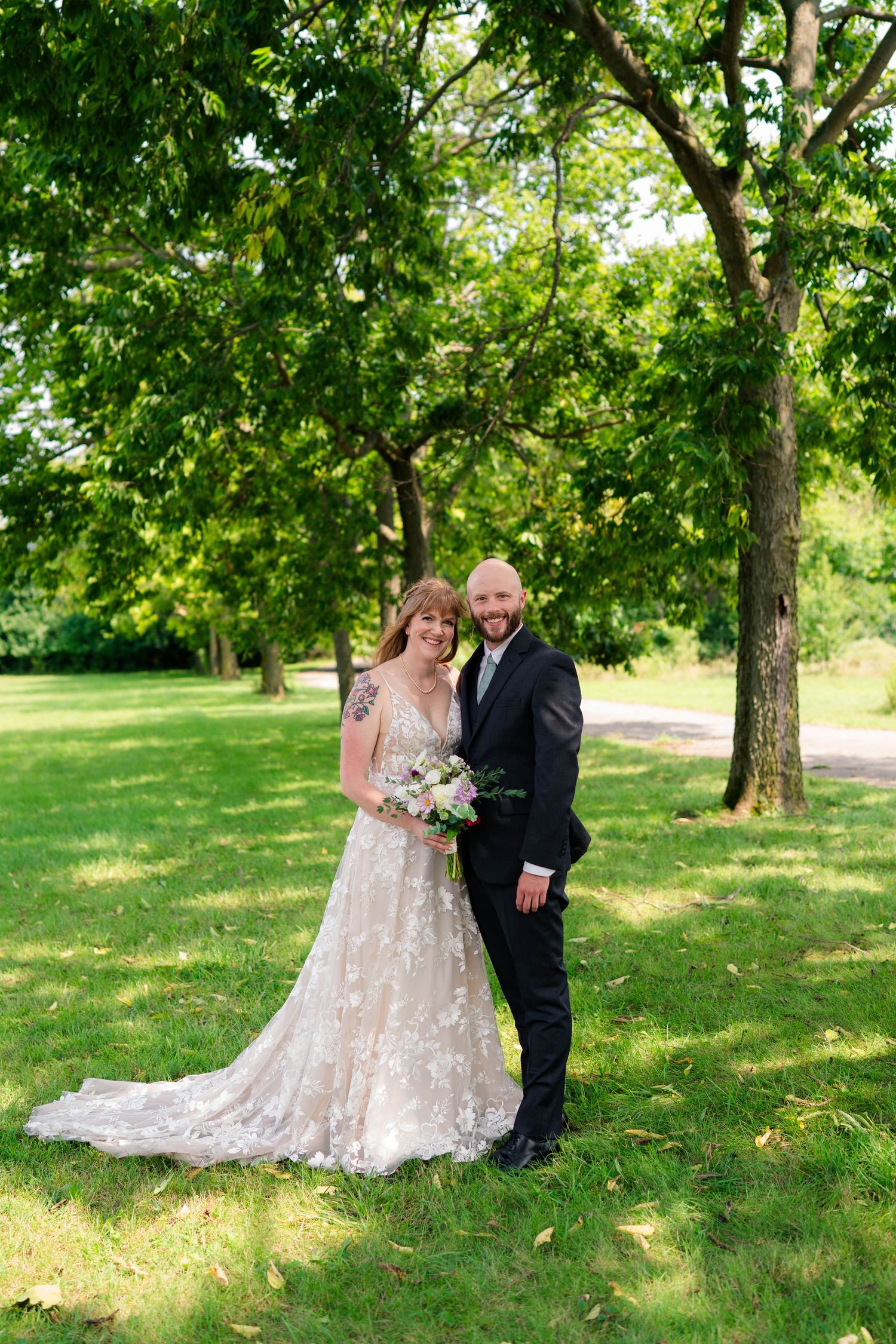 Smiling bride and bald groom pose together in a lush green park, bride holding a floral bouquet.
