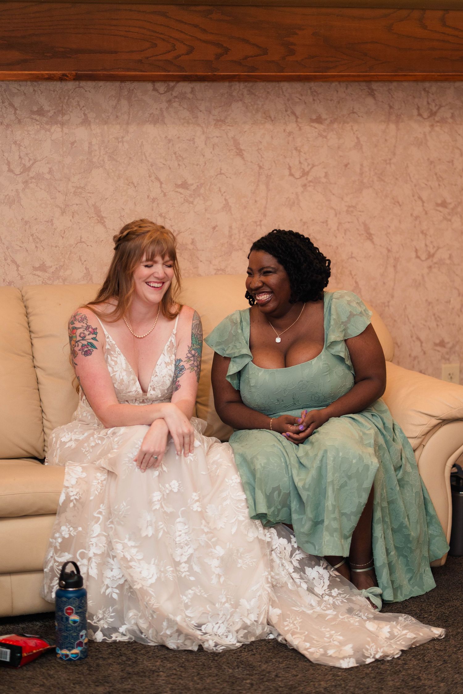 Bride in lace gown laughing with bridesmaid in green dress on cream sofa at wedding.