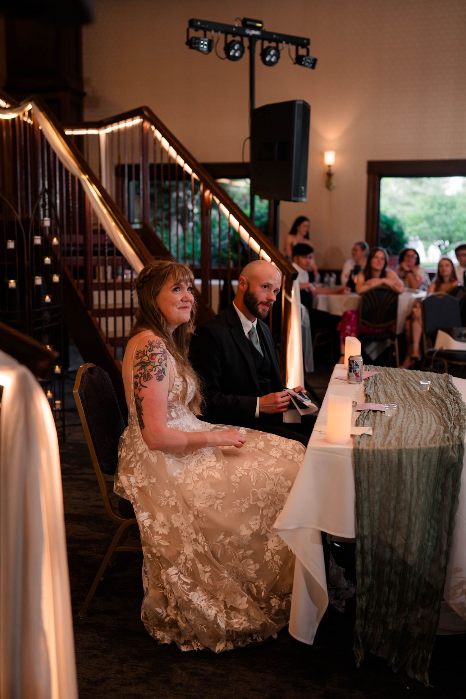 Bride and groom seated at sweetheart table listening to speeches during wedding reception.