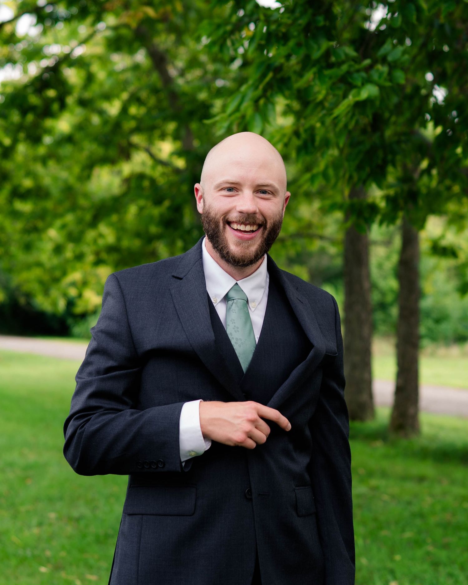 A smiling bald groom in a dark suit with sage green tie poses outdoors among green trees.