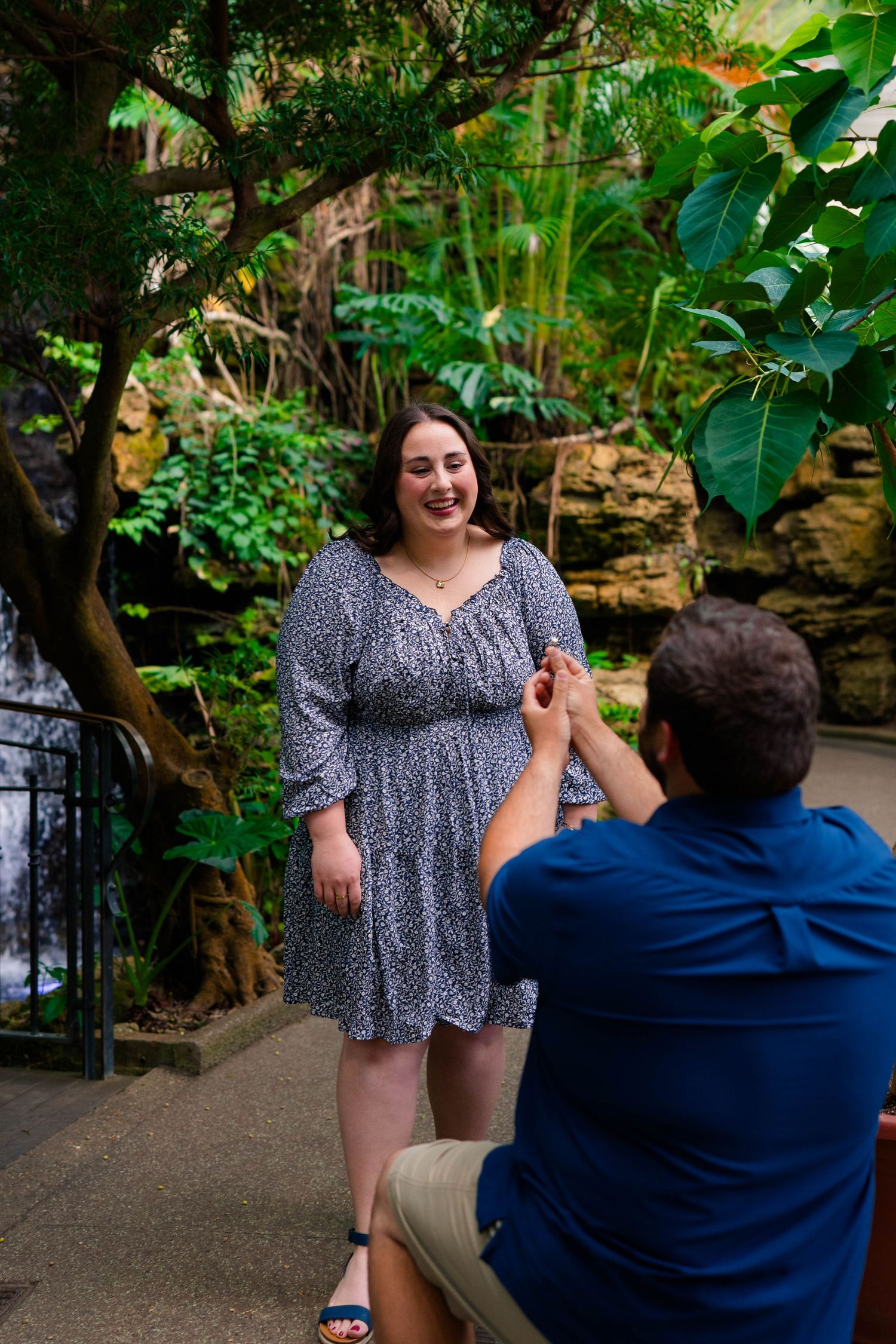 Man kneeling on one knee proposing to a smiling woman in a floral dress in a lush tropical garden.